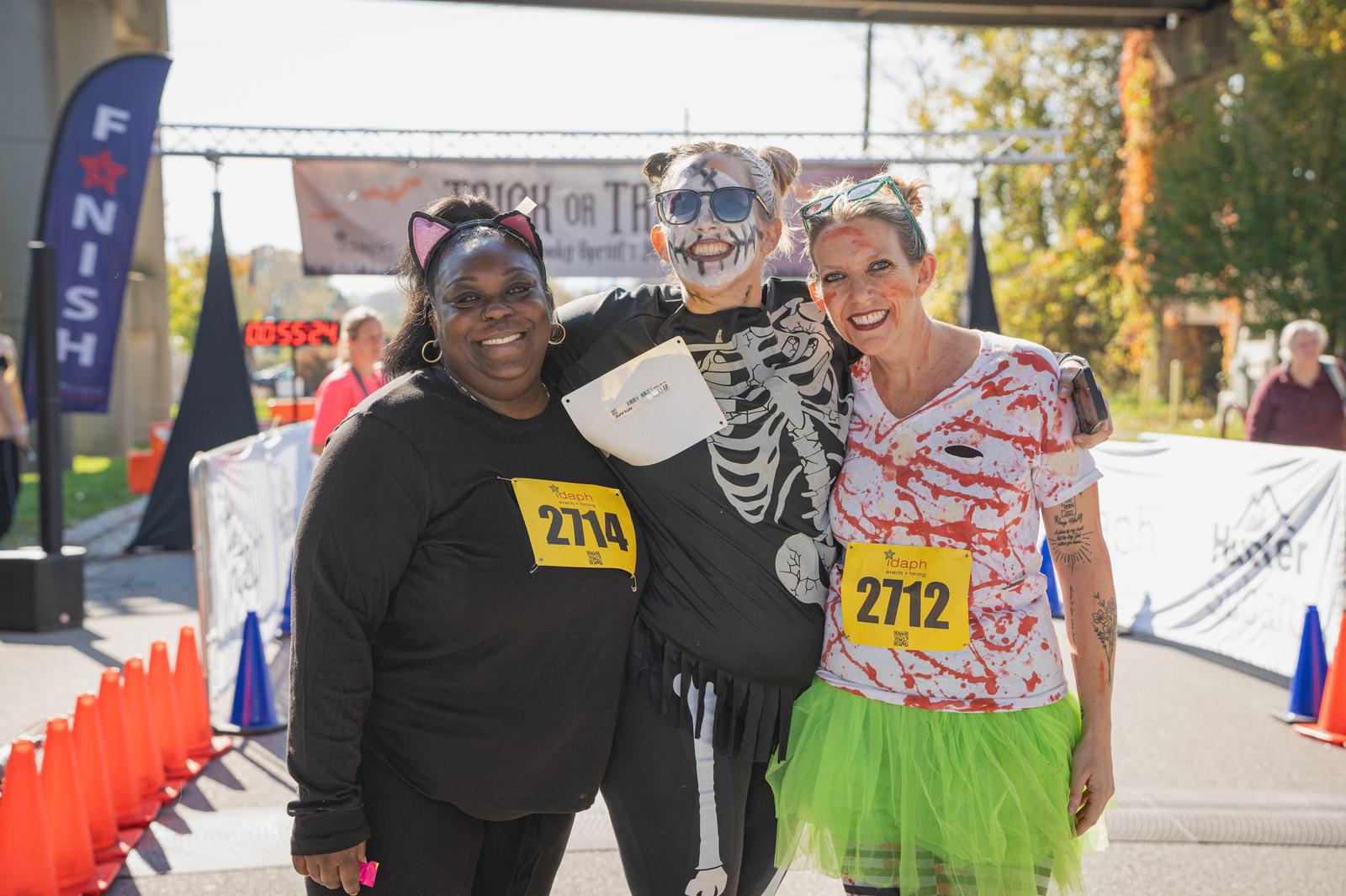 Three smiling women in Halloween costumes pose at a race finish line. One is dressed as a cat, one as a skeleton, and one in a green tutu with a bloody shirt. All wear race bibs and enjoy a sunny day outdoors.