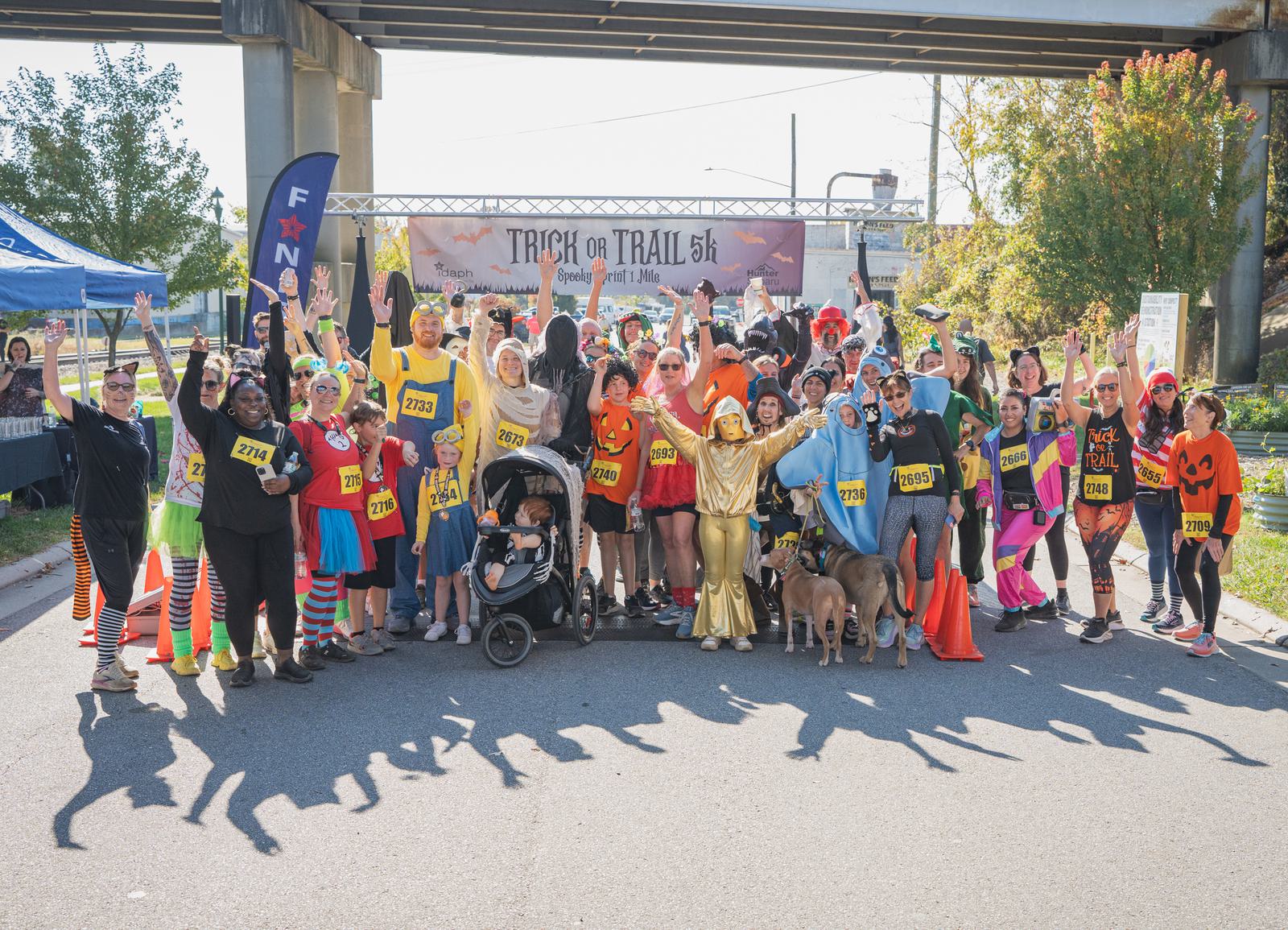 A large group of people in colorful costumes pose and raise their arms at the starting line of a "Trick or Trail 5K" race, with a banner and festive decorations visible in the background.