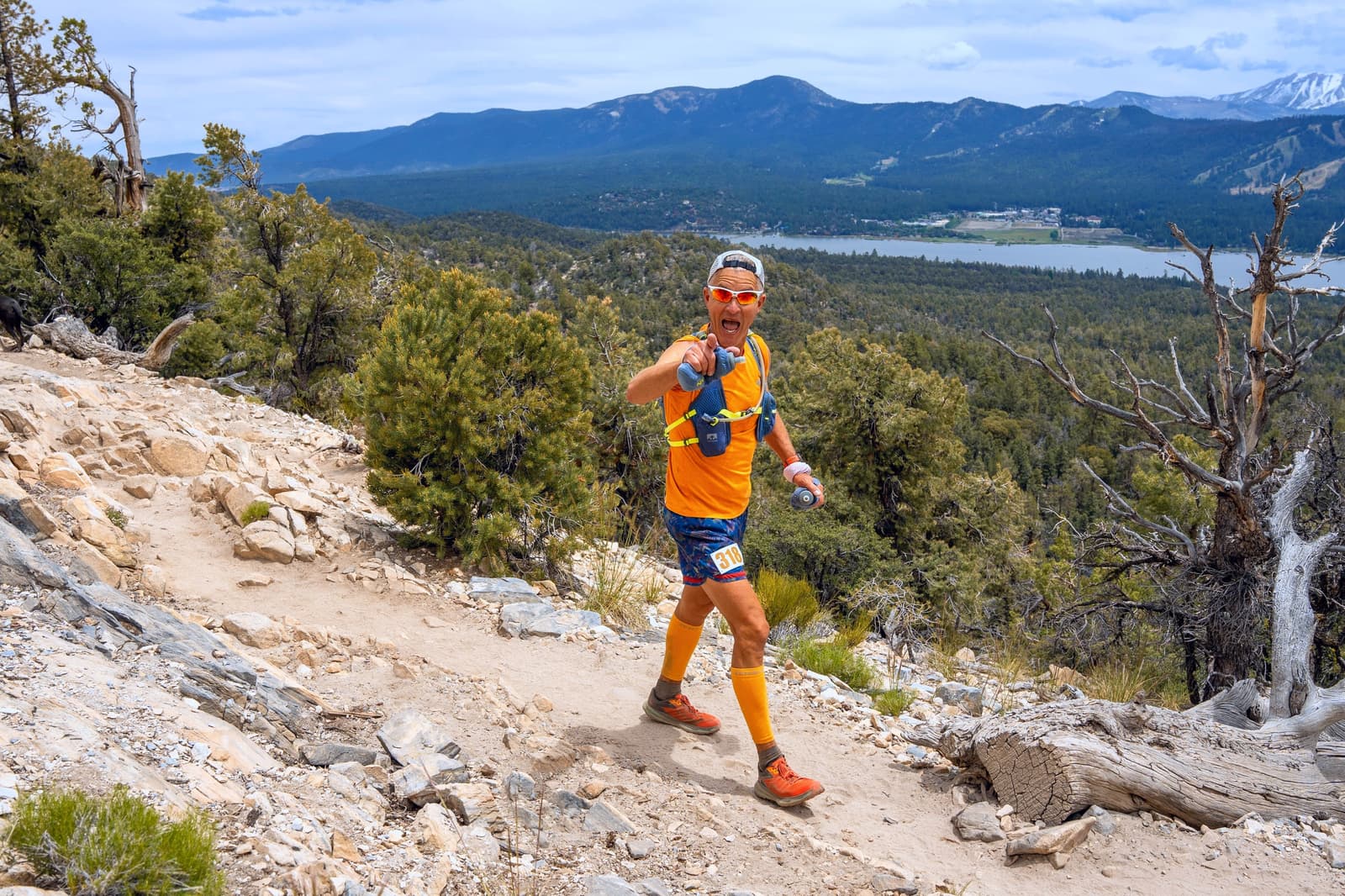 A person in bright orange athletic gear and sunglasses runs on a rocky mountain trail. They give a thumbs-up gesture. The background features a scenic view of mountains and a lake under a partly cloudy sky.