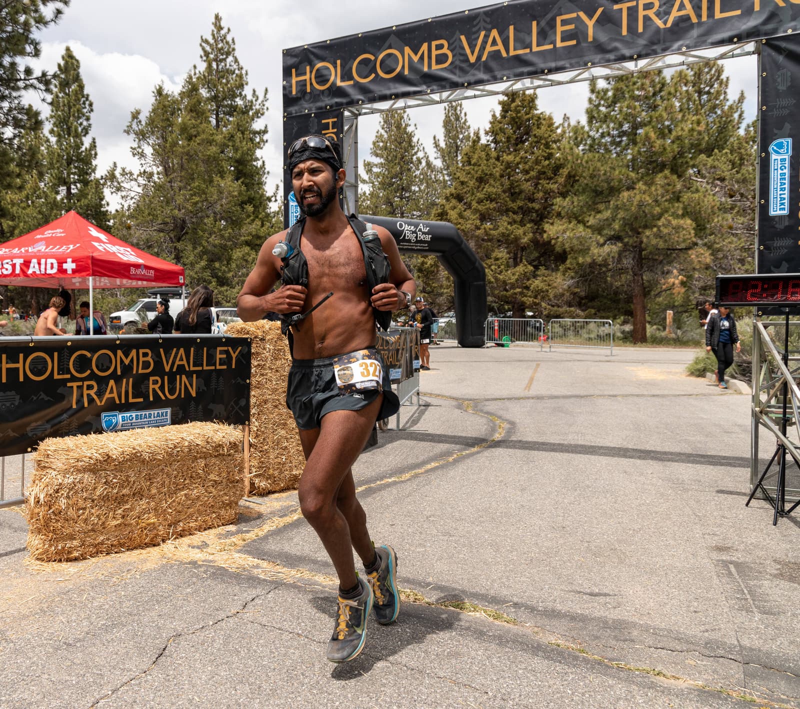 A runner crosses the finish line at the Holcomb Valley Trail Run. He is shirtless, wearing a cap, shorts, and a hydration pack. The finish line banner and trees are visible in the background. A digital clock shows his finishing time.