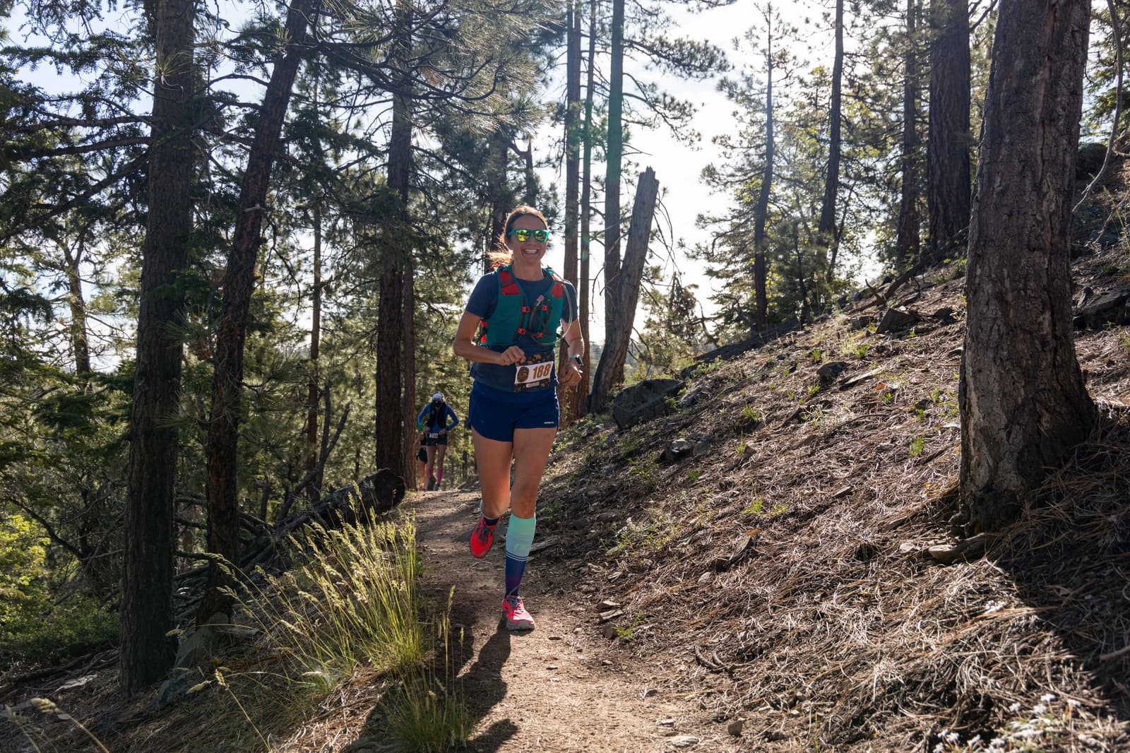 A person running on a dirt trail through a forested area. They are wearing sunglasses, a hydration vest, and a race bib. The sun is shining through the trees, casting dappled shadows on the ground. Another runner is visible in the background.