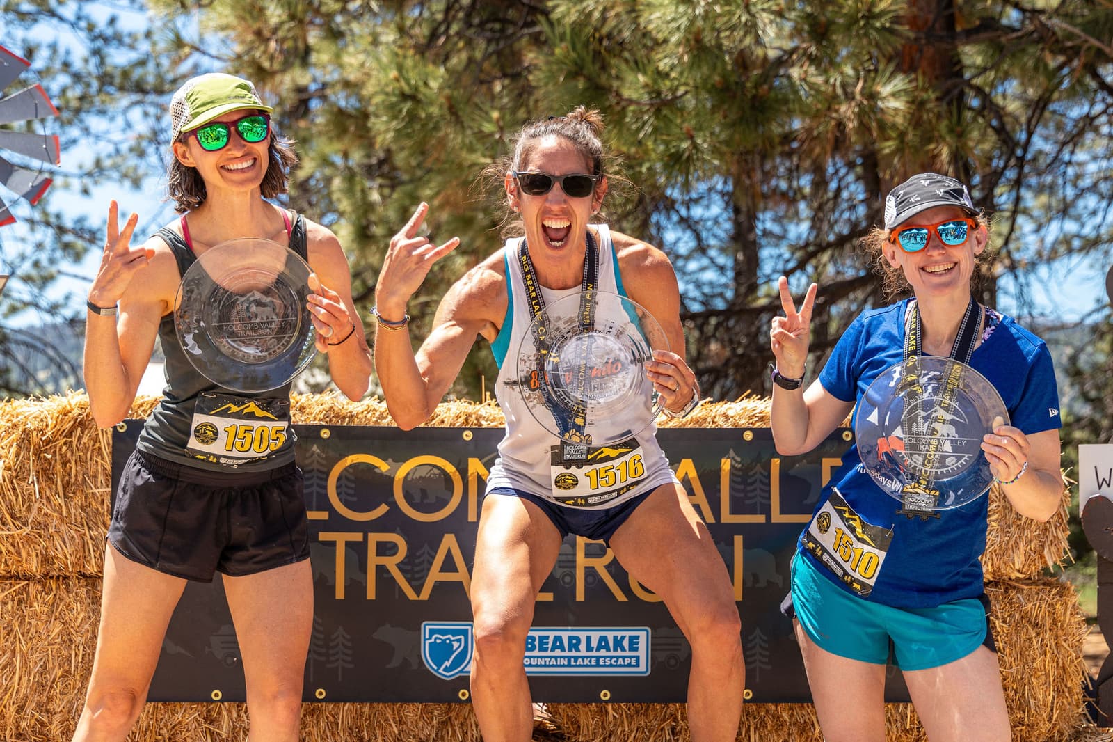 Three women celebrate on a podium at a trail run event, each holding a finisher's plaque. They are smiling and making peace signs. They wear running gear and sunglasses, with trees and a banner in the background.