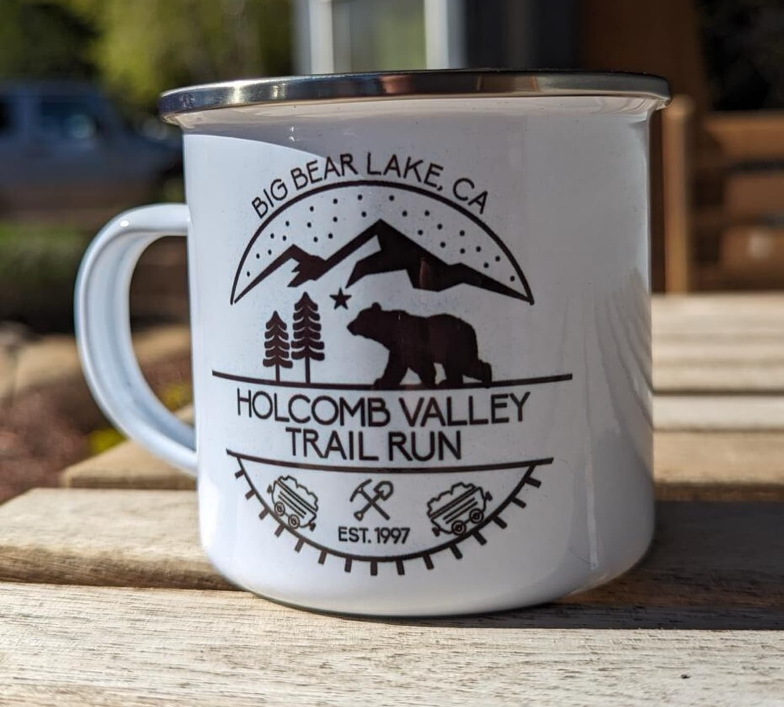 A white metal mug with a logo for "Holcomb Valley Trail Run" featuring a bear, trees, and mountains. Text includes "Big Bear Lake, CA" and "Est. 1997." The mug is on a wooden surface with a blurred outdoor background.