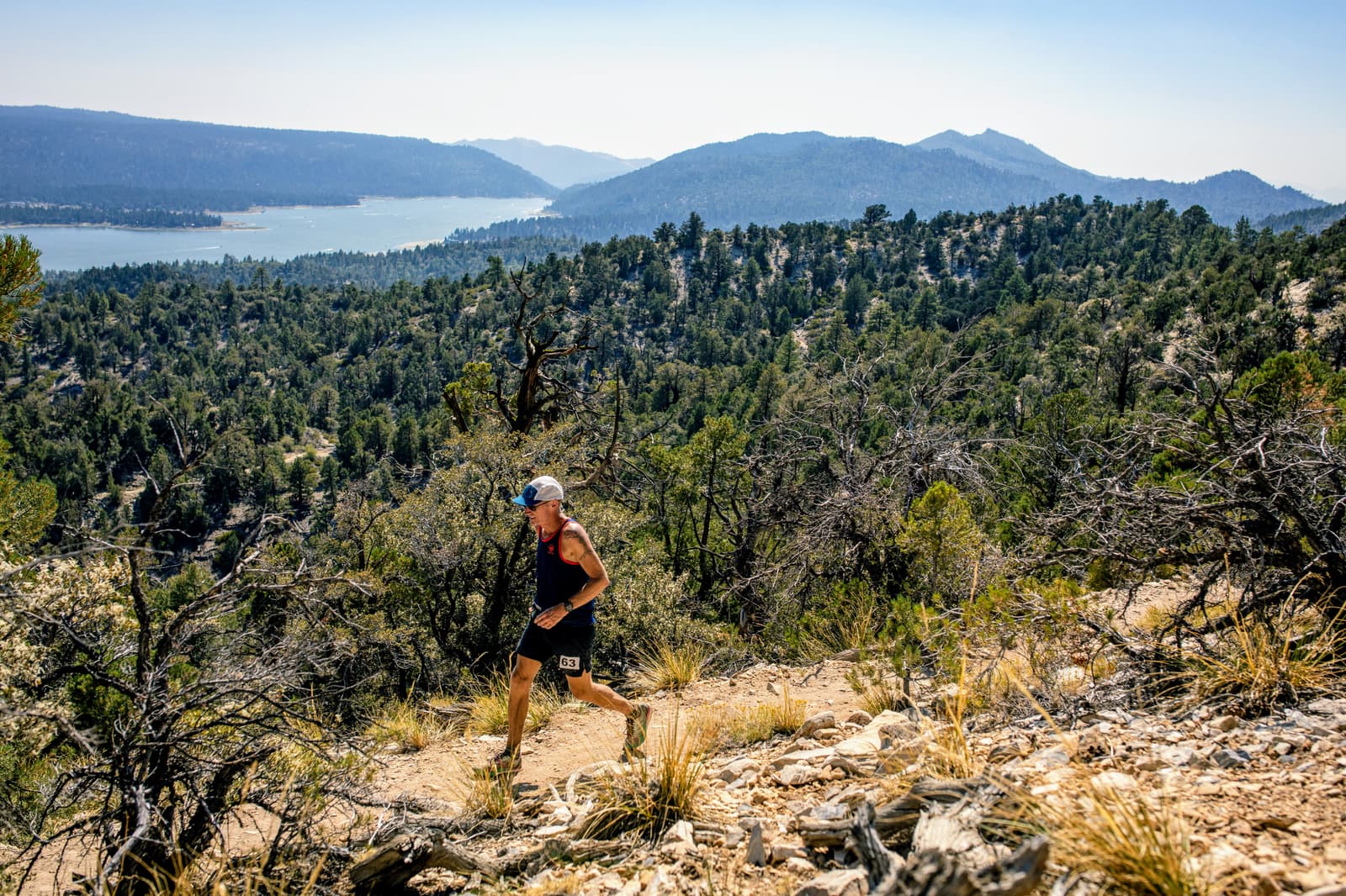 A person is running on a rocky trail in a mountainous area. They wear a cap, tank top, shorts, and a race bib. In the background, there are lush, forested hills and a body of water with distant mountains under a clear sky.