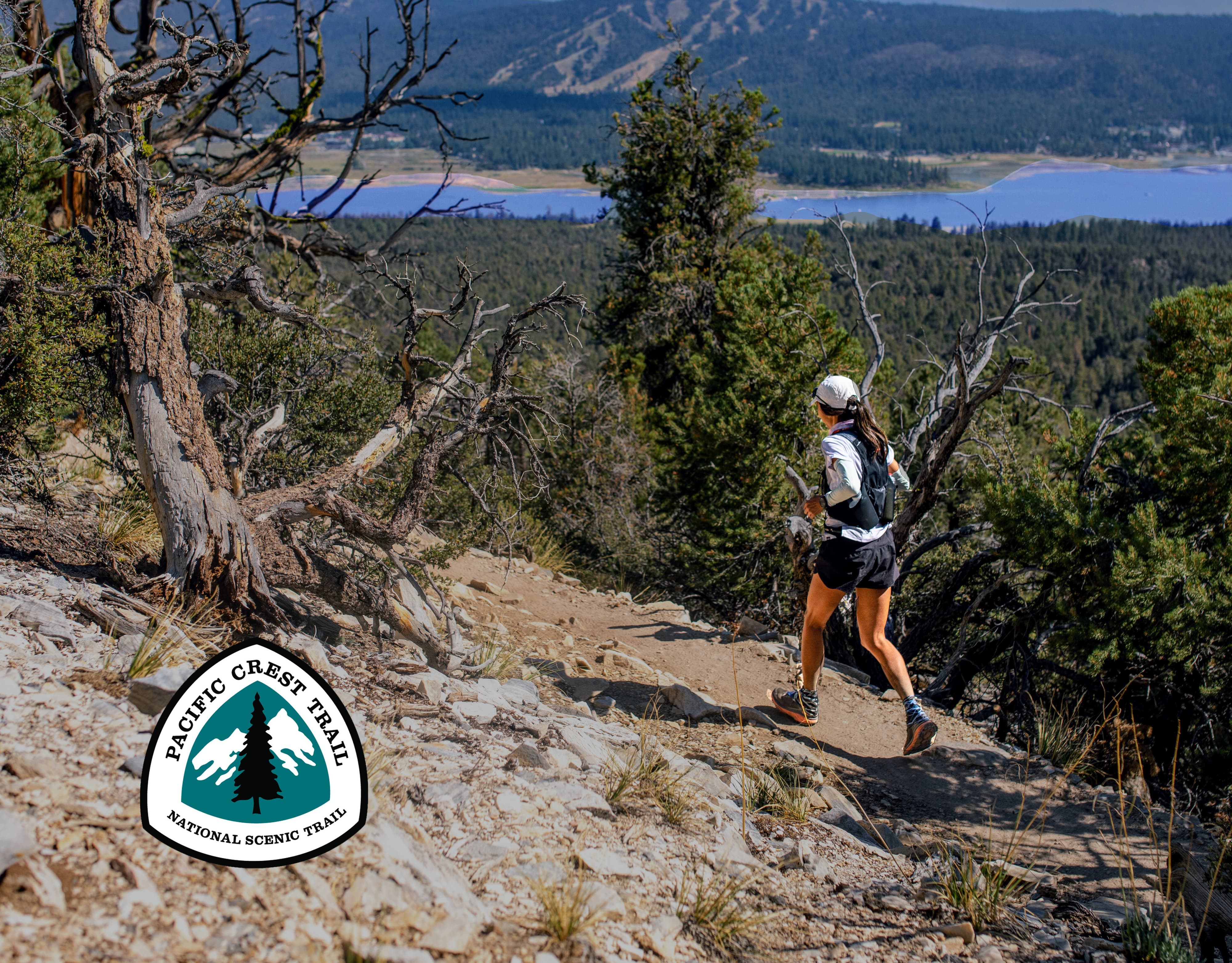 A person is trail running on a rocky path surrounded by trees and mountains. They are wearing a cap, backpack, and athletic gear. A Pacific Crest Trail logo is in the foreground. A distant lake and forested hills are visible under a clear blue sky.