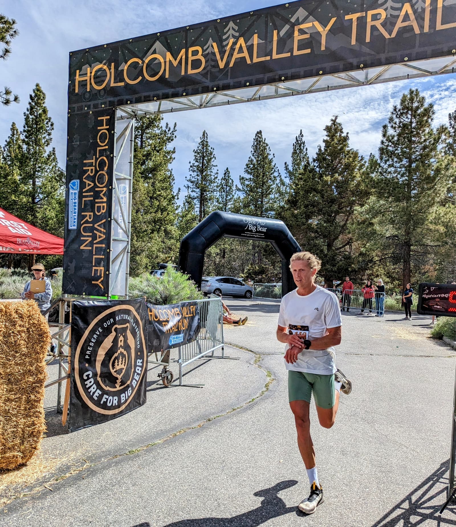 A runner in a white shirt and green shorts approaches the finish line of the Holcomb Valley Trail Run. The archway above reads "Holcomb Valley Trail Run." Spectators and pine trees are visible in the background.