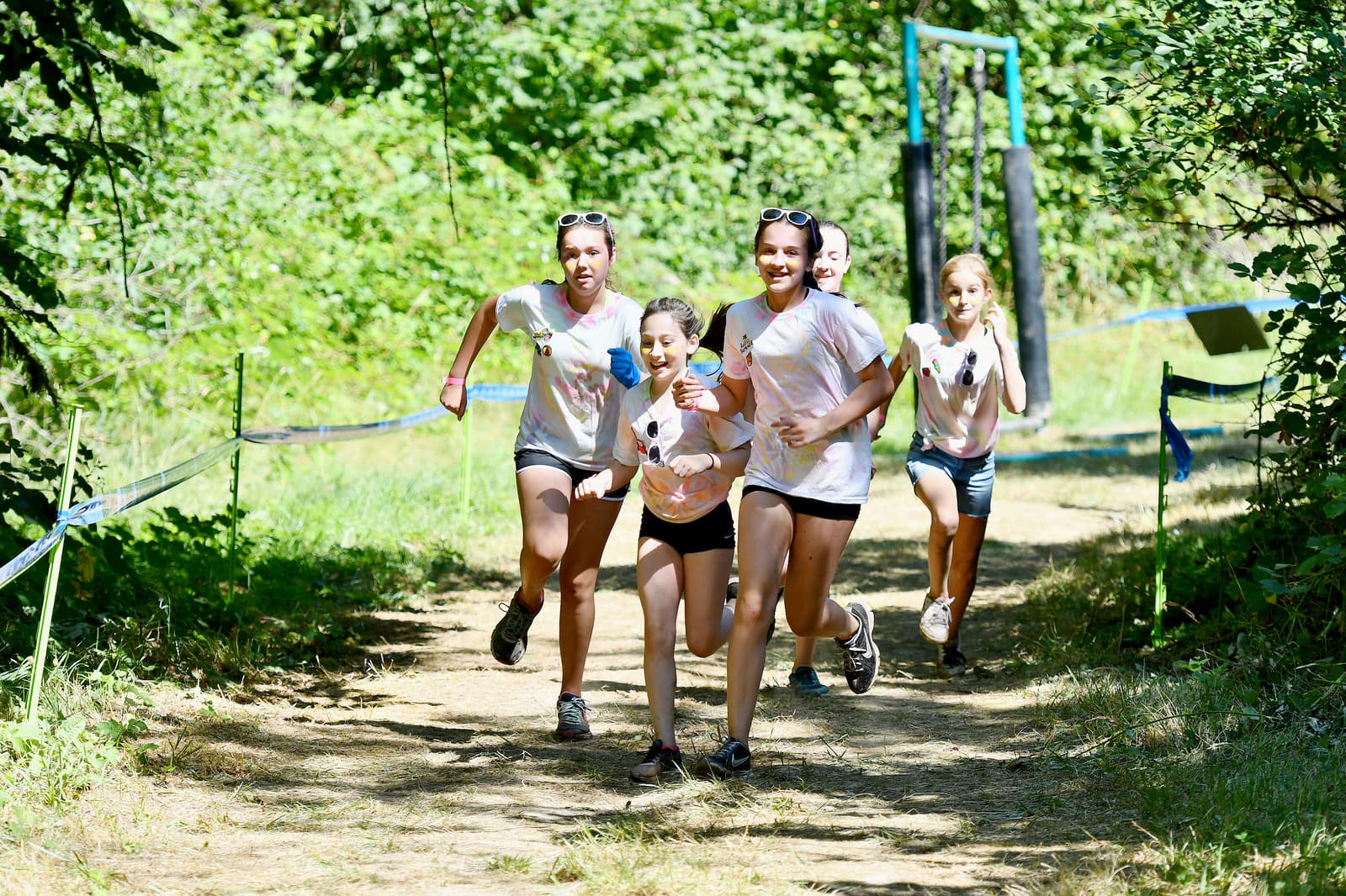 Five smiling girls running on a forest trail during a sunny day, wearing matching white shirts with colorful designs. They hold water bottles and seem to be participating in a fun outdoor event or race. Trees and greenery surround the path.