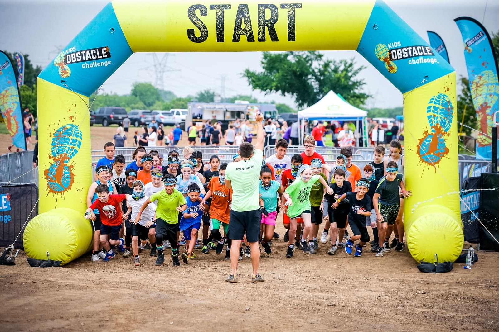 Children dressed in colorful athletic gear excitedly begin an obstacle challenge race, passing under a yellow "Start" banner. An adult in front encourages them. Background includes spectators and a white tent.