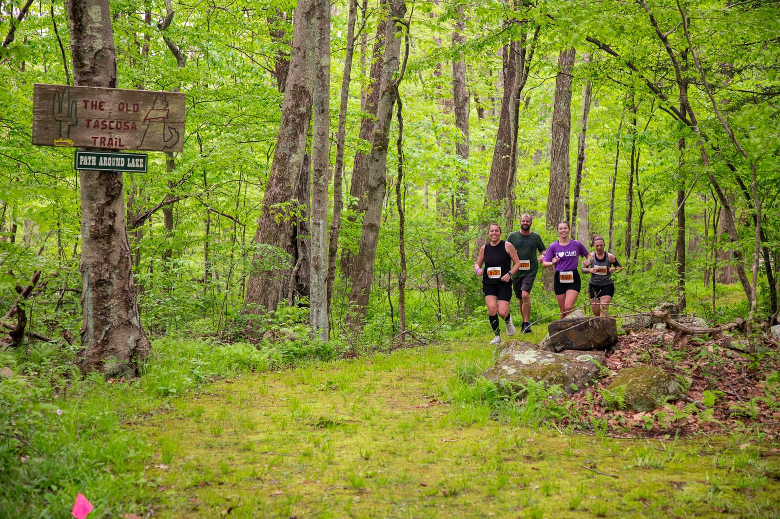 Four people are running on a lush forest trail. A wooden sign on a tree reads "The Old Tascosa Trail" and "Palo Duro Rail." The ground is covered in greenery and fallen leaves, creating a vibrant natural setting.
