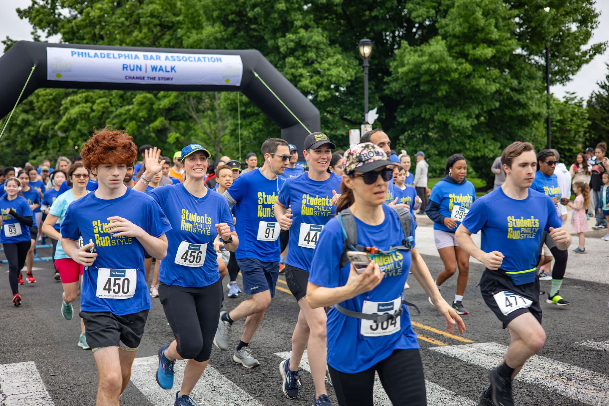 A group of runners in blue "Students Run Philly Style" shirts participate in a race. They are running on a road under a black archway that reads "Philadelphia Bar Association Run/Walk." Green trees line the background.