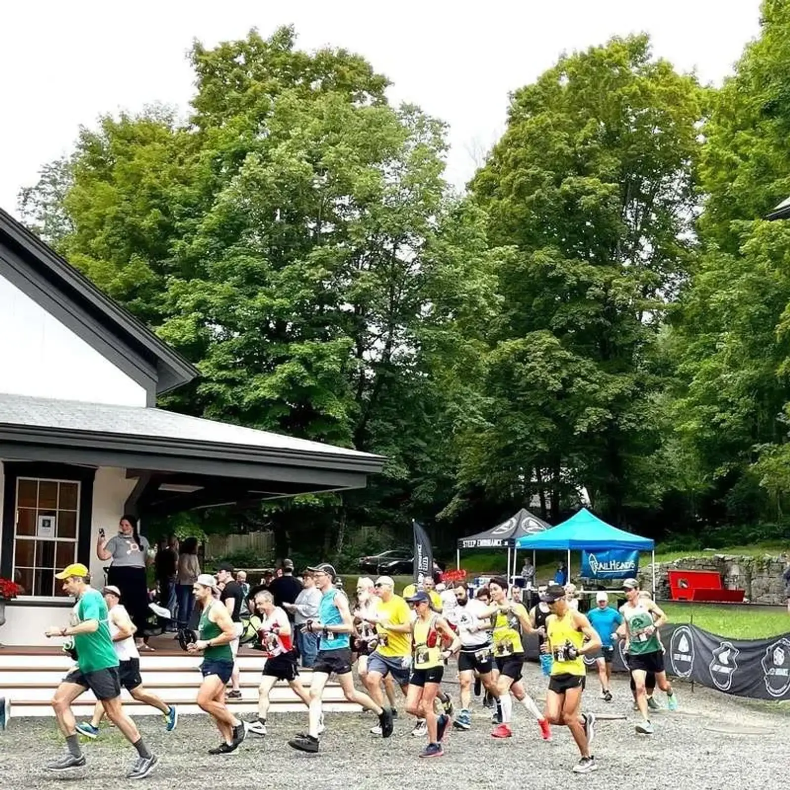 A group of runners in athletic gear is competing in a race, passing by a building with onlookers cheering. The background is filled with lush green trees and a blue canopy tent. The atmosphere is lively and energetic.
