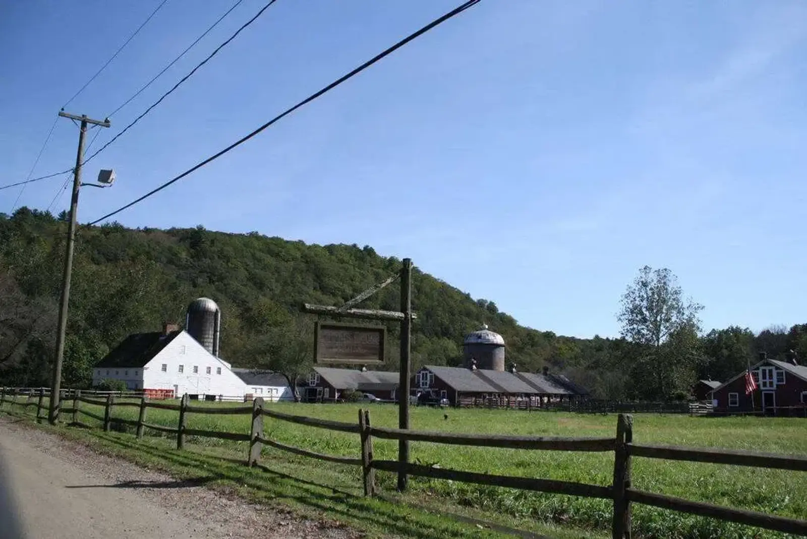 Rural scene with a dirt road beside a wooden fence, leading past a farm with multiple buildings, including a white barn and silos. Green grass covers the fields, and a hill with dense trees is in the background under a clear blue sky.