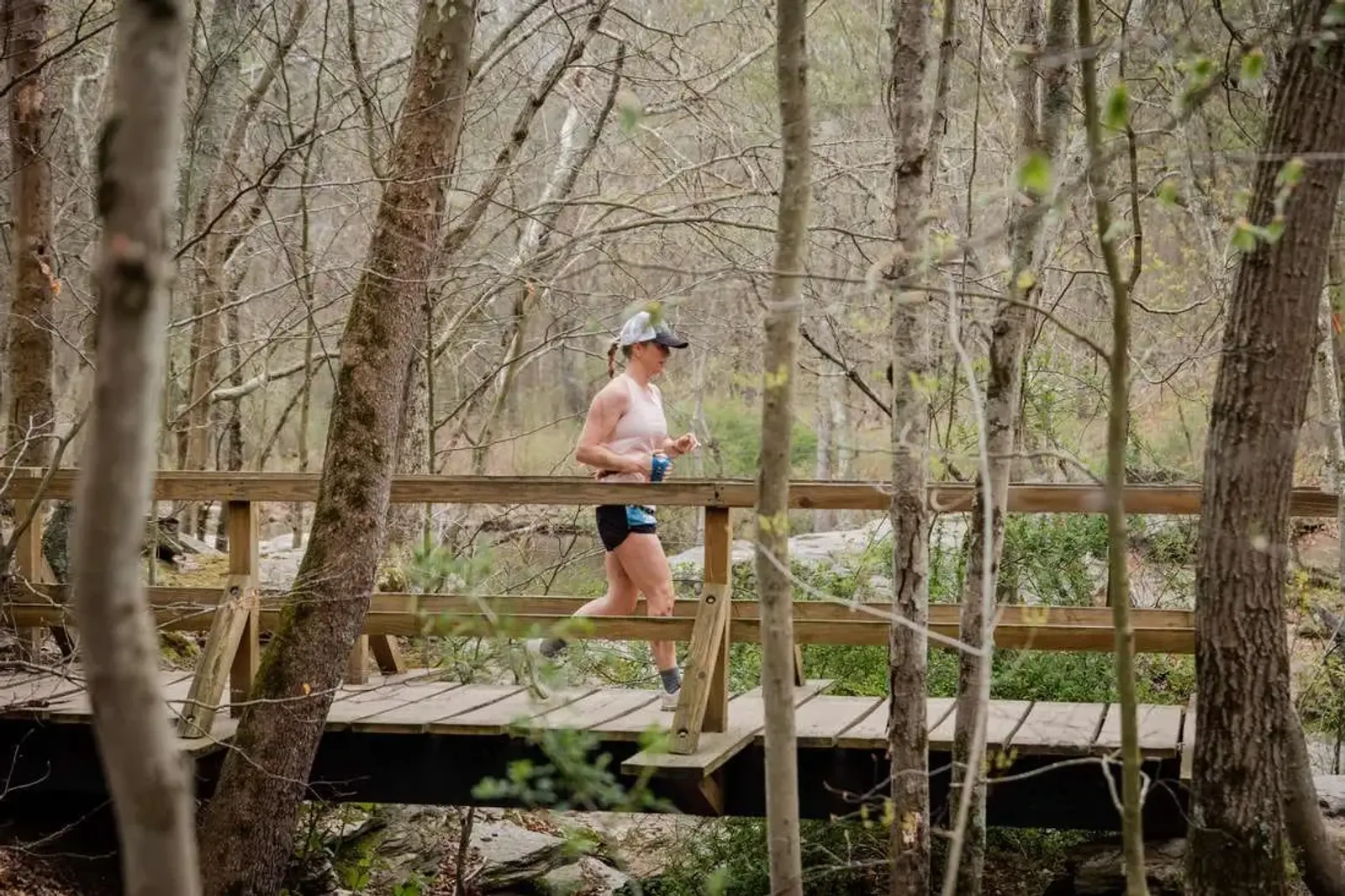 A person runs across a wooden bridge in a forest. They wear a tank top, shorts, and a cap, holding a drink. Bare trees and greenery surround the bridge, suggesting early spring or fall.