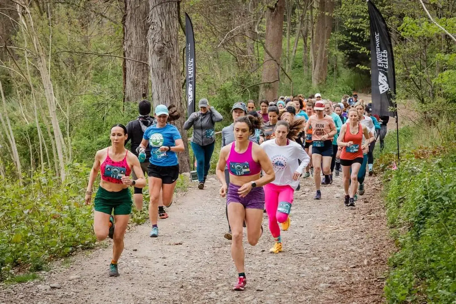 A group of runners on a forest trail, participating in a race. Most wear colorful athletic clothing, with some in shorts and tank tops. The path is surrounded by trees and greenery, and there are flags marking the race route.
