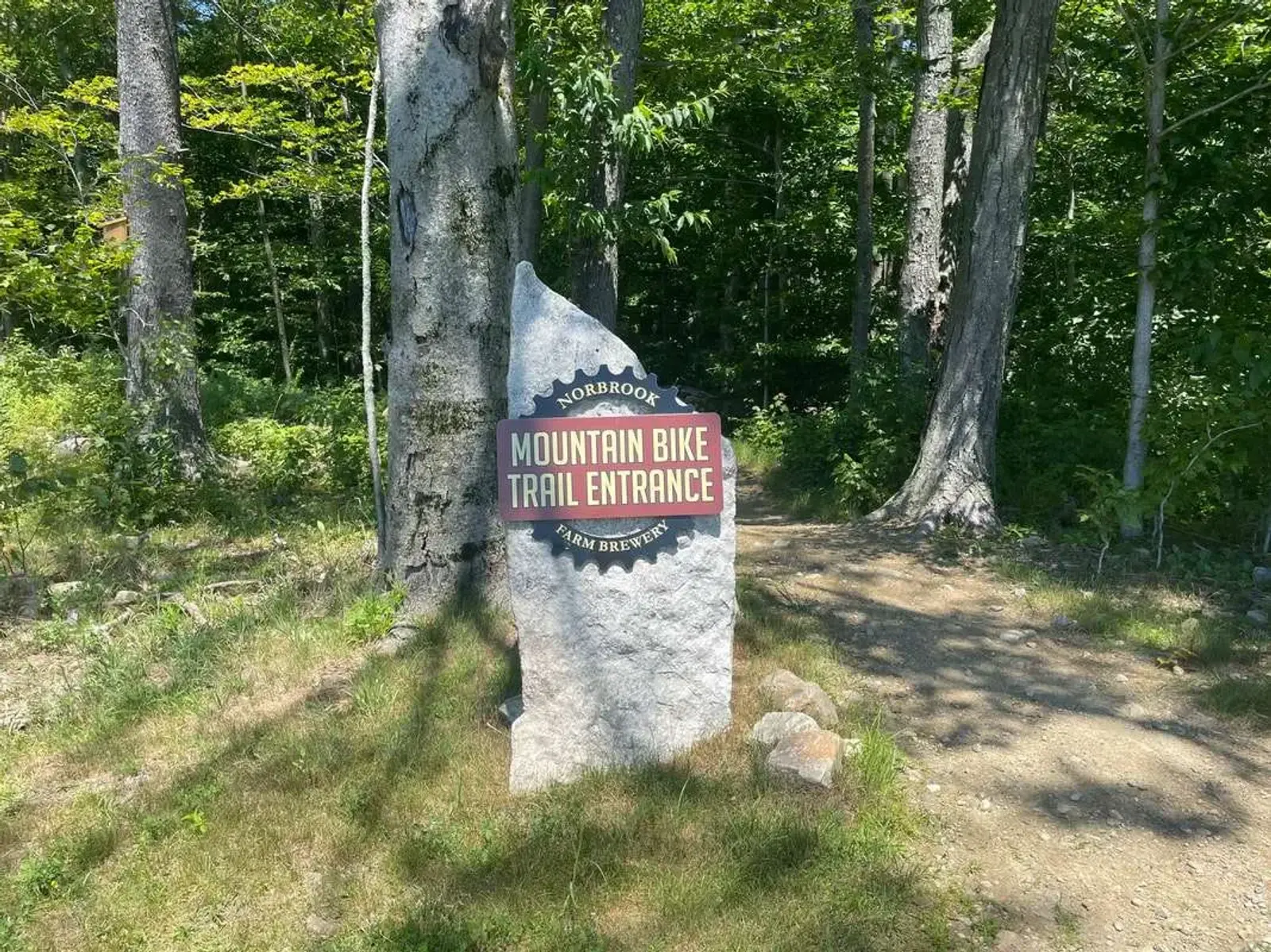 A stone marker under trees reads "Noorbrook Mountain Bike Trail Entrance, Sam Brewster" in the sunlight, marking the start of a forested trail.