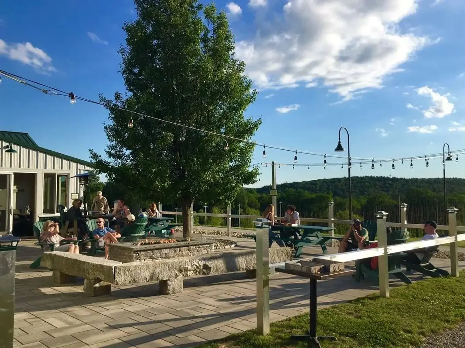 People sit at picnic tables on a patio with string lights, a tree, and a fire pit. The sky is blue with some clouds, and green hills are visible in the background. A small building is on the left.