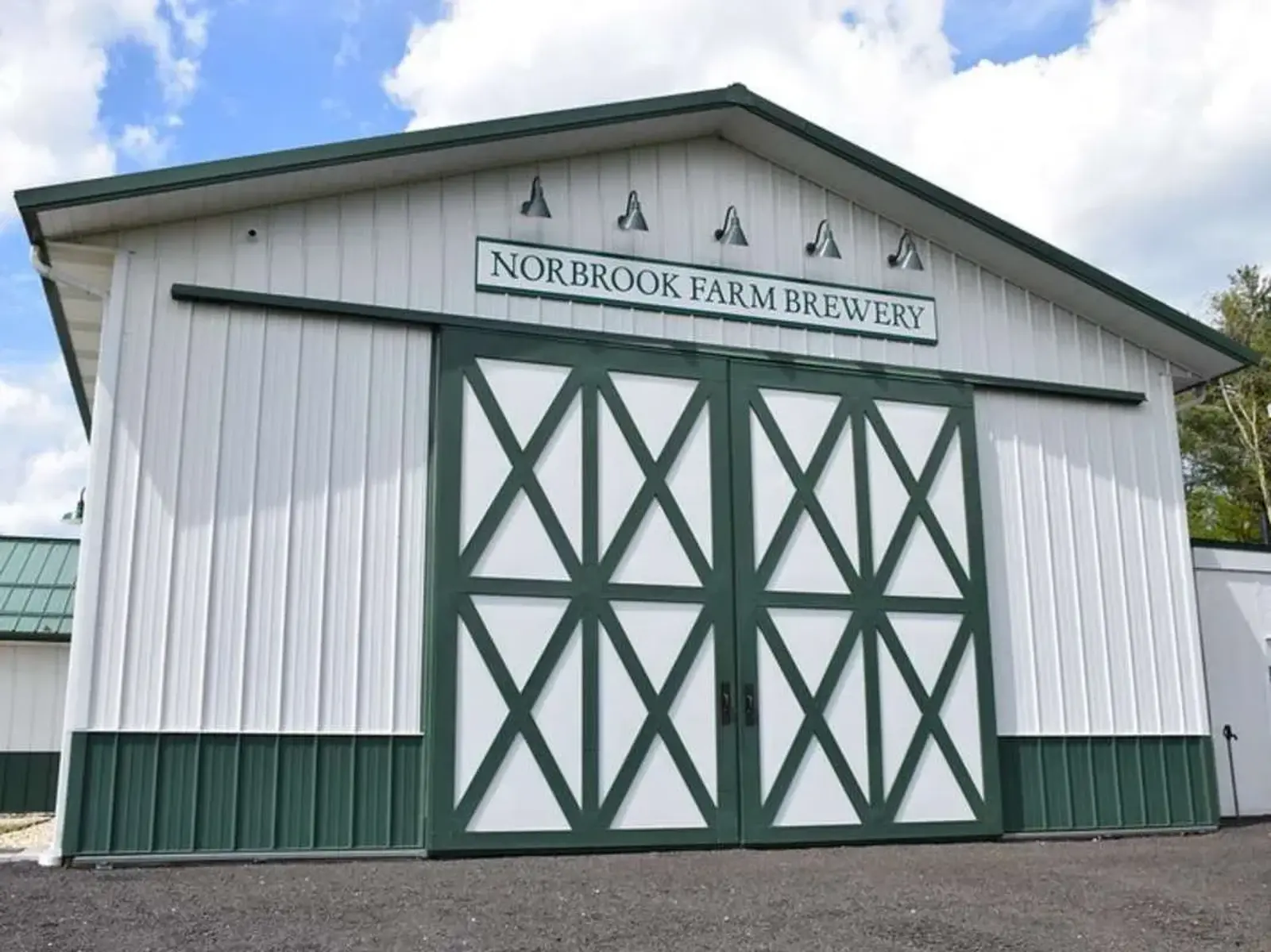 A white barn-style building with green trim and large double doors reads "Norbrook Farm Brewery" above the entrance, under a partly cloudy sky.