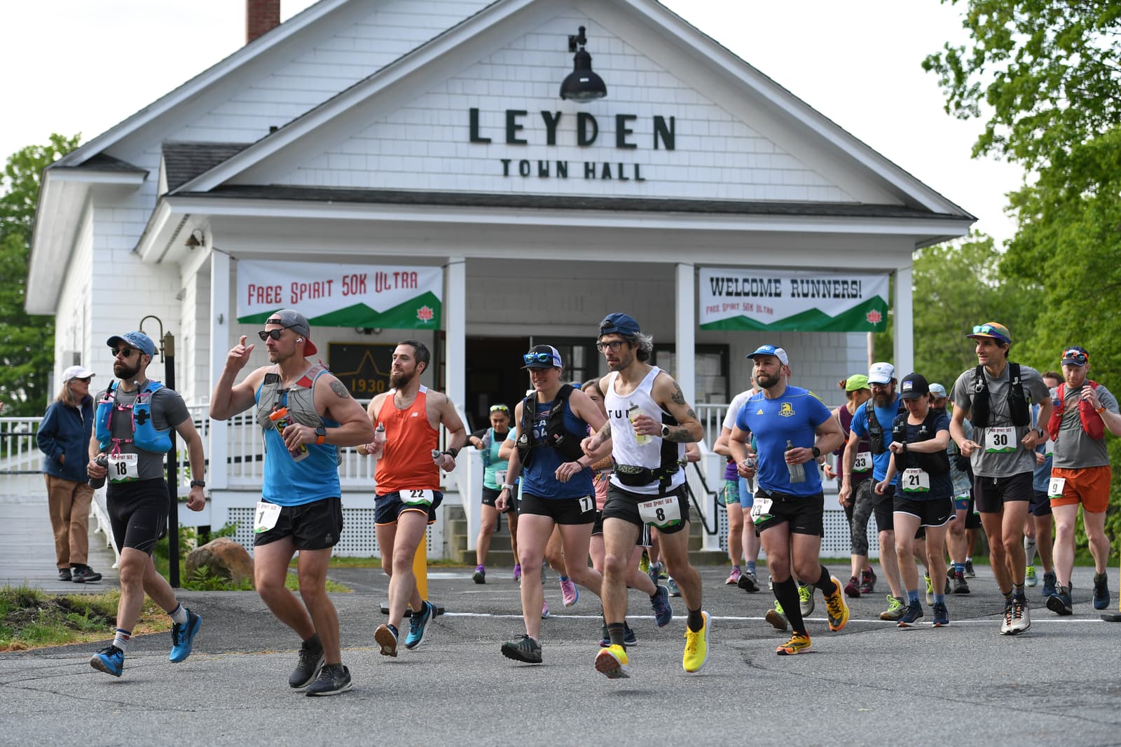 A group of runners wearing race bibs and athletic gear start a race in front of Leyden Town Hall, with signs welcoming runners and reading "Free Spirit 50K Ultra" visible on the building.