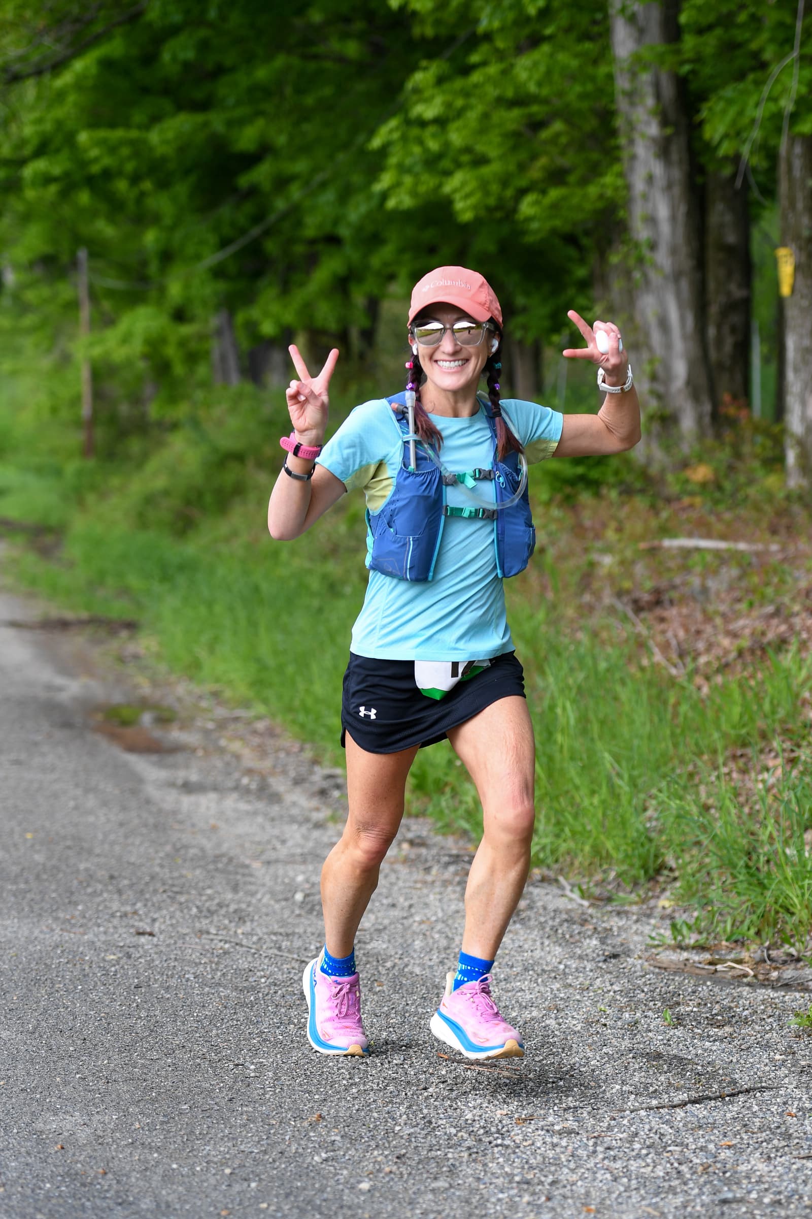 A woman running outdoors on a paved road, smiling and making peace signs with both hands. She wears a pink cap, sunglasses, light blue shirt, black shorts, running vest, and pink running shoes. Green trees line the background.