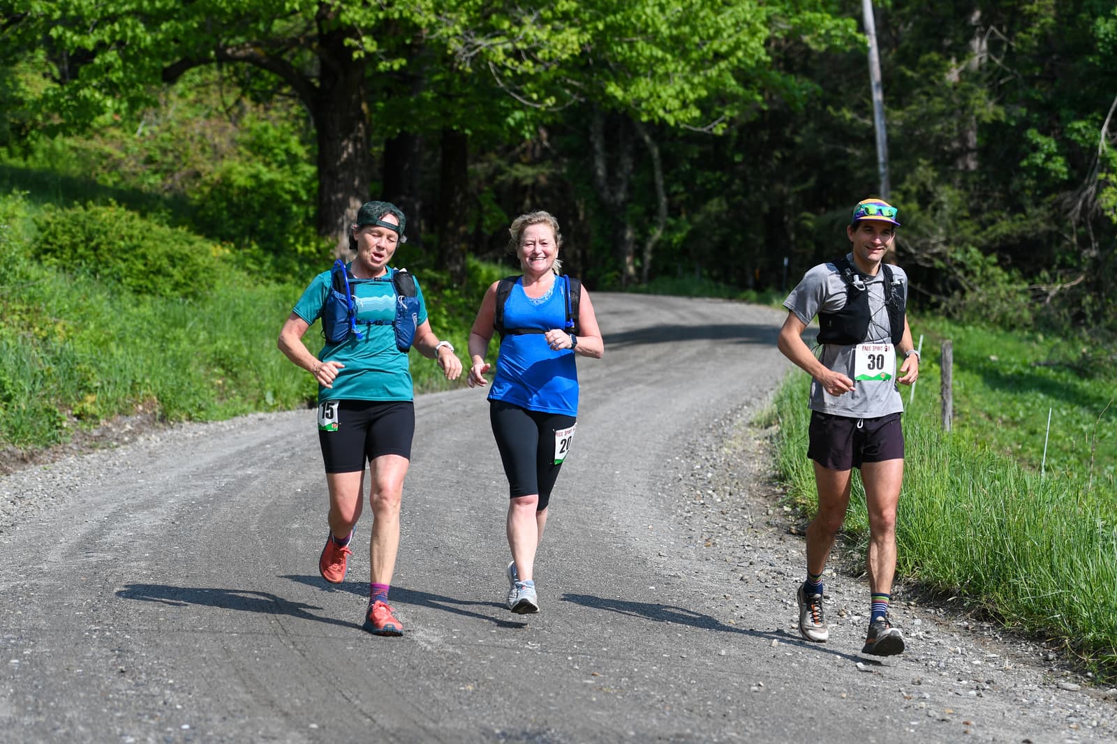 Three runners are jogging on a gravel road surrounded by greenery, wearing athletic gear and race bibs, smiling and enjoying the run on a sunny day.