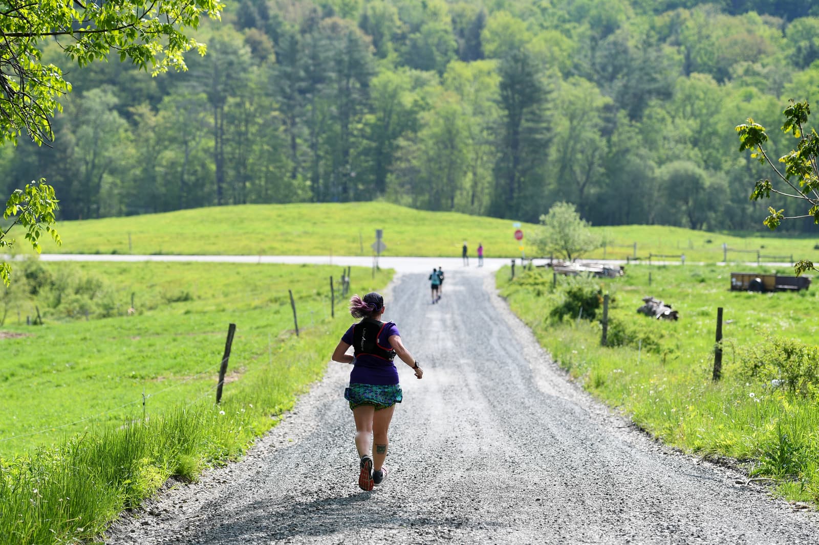 A person runs on a gravel road through a lush, green countryside with trees and grass. Other runners are visible in the distance under a clear, sunny sky.