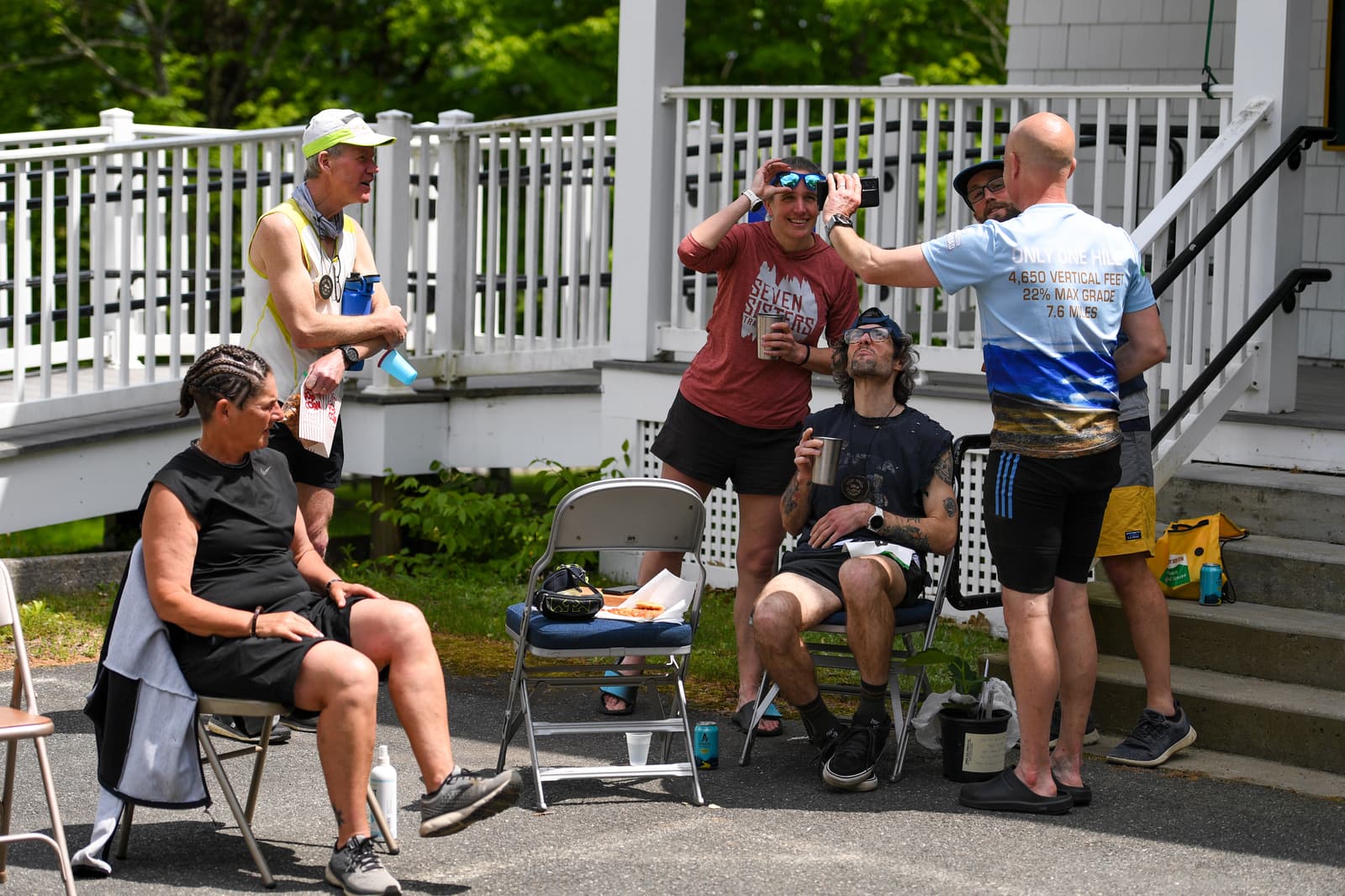 A group of runners take a break near a white porch. Some sit on folding chairs while others stand, talking and laughing. One person applies something to another’s face as others relax and chat in casual athletic gear.