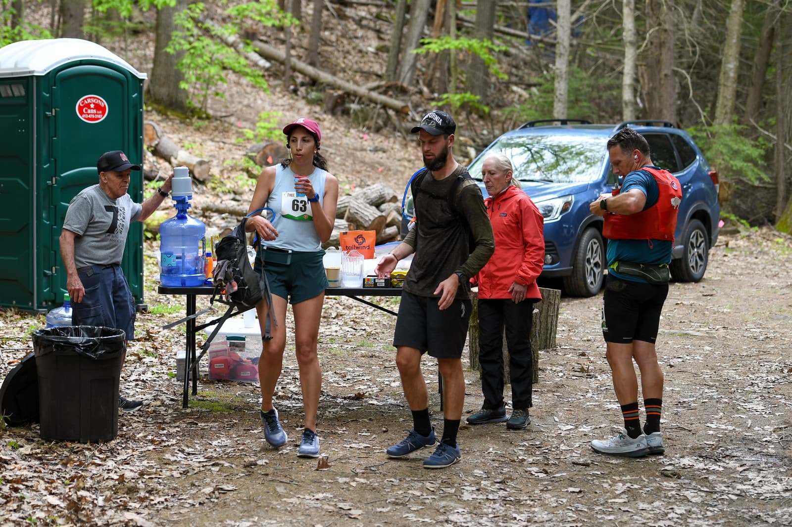 Runners stop at a trail-side aid station with snacks, water, and volunteers in a wooded area. A portable toilet and car are in the background. One runner wears bib 63 and adjusts her cap.