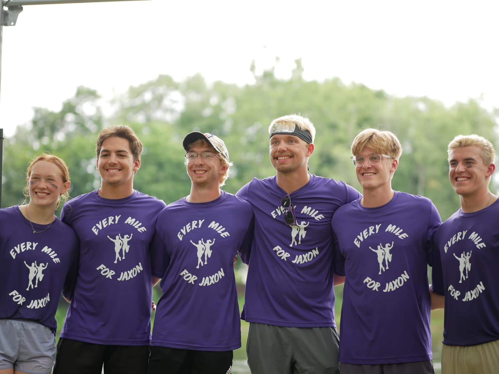 Six smiling young adults stand side by side, wearing matching purple shirts that say "Every Mile For Jaxon" with a graphic of three runners. Trees and greenery are visible in the blurred background.