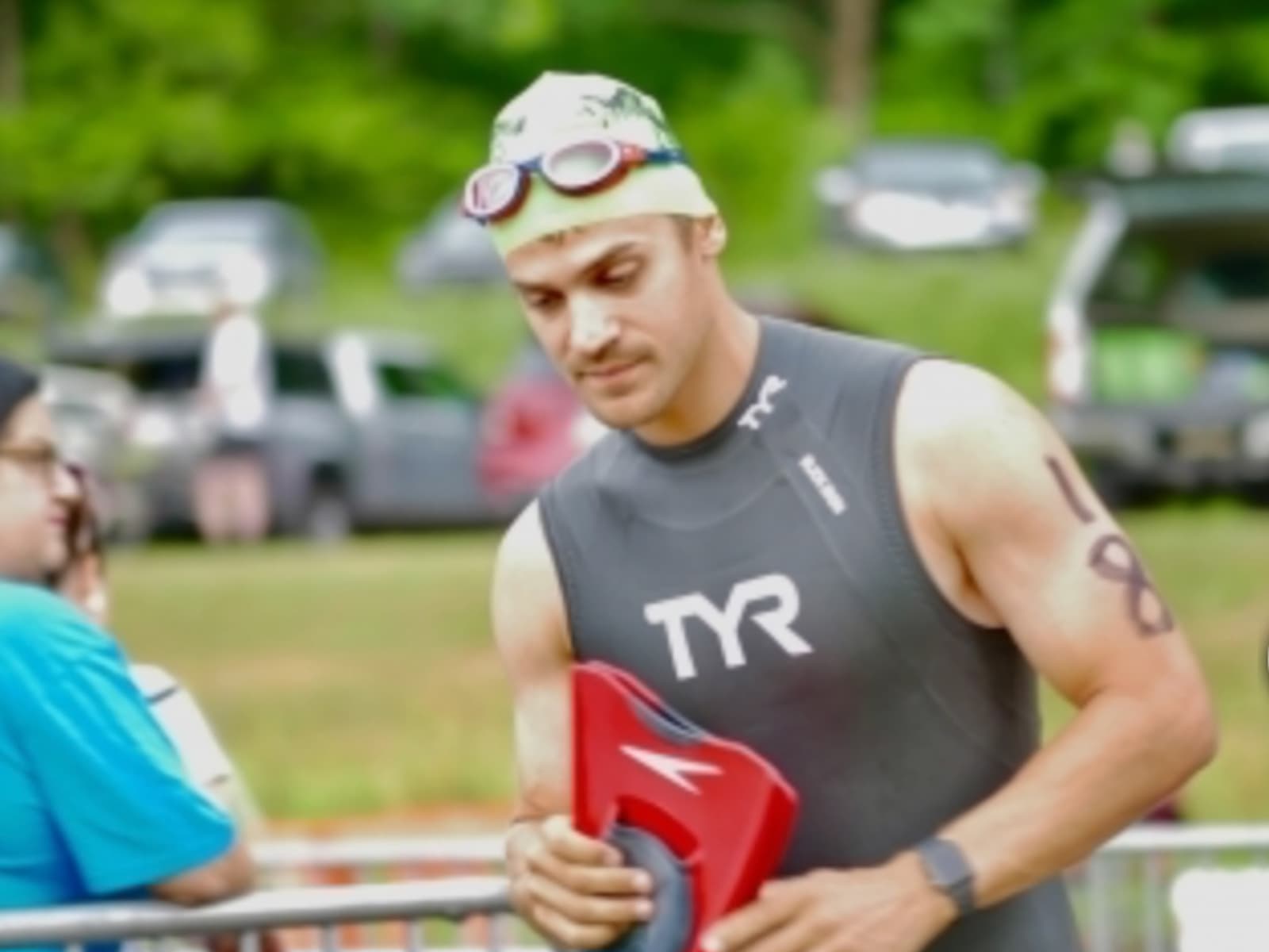 A man wearing a swim cap, goggles, and a black TYR wetsuit stands outdoors at a sporting event, holding red swim paddles. His upper arm features the numbers "18" written with a marker. There are blurred people and cars in the background.