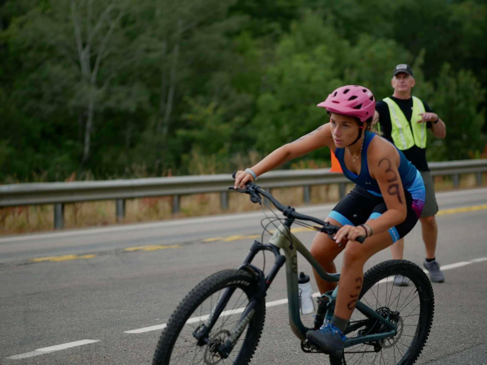 A cyclist wearing a pink helmet and race numbers on their arms rides on a road, focused ahead. A person in a yellow vest stands in the background near a guardrail and trees.