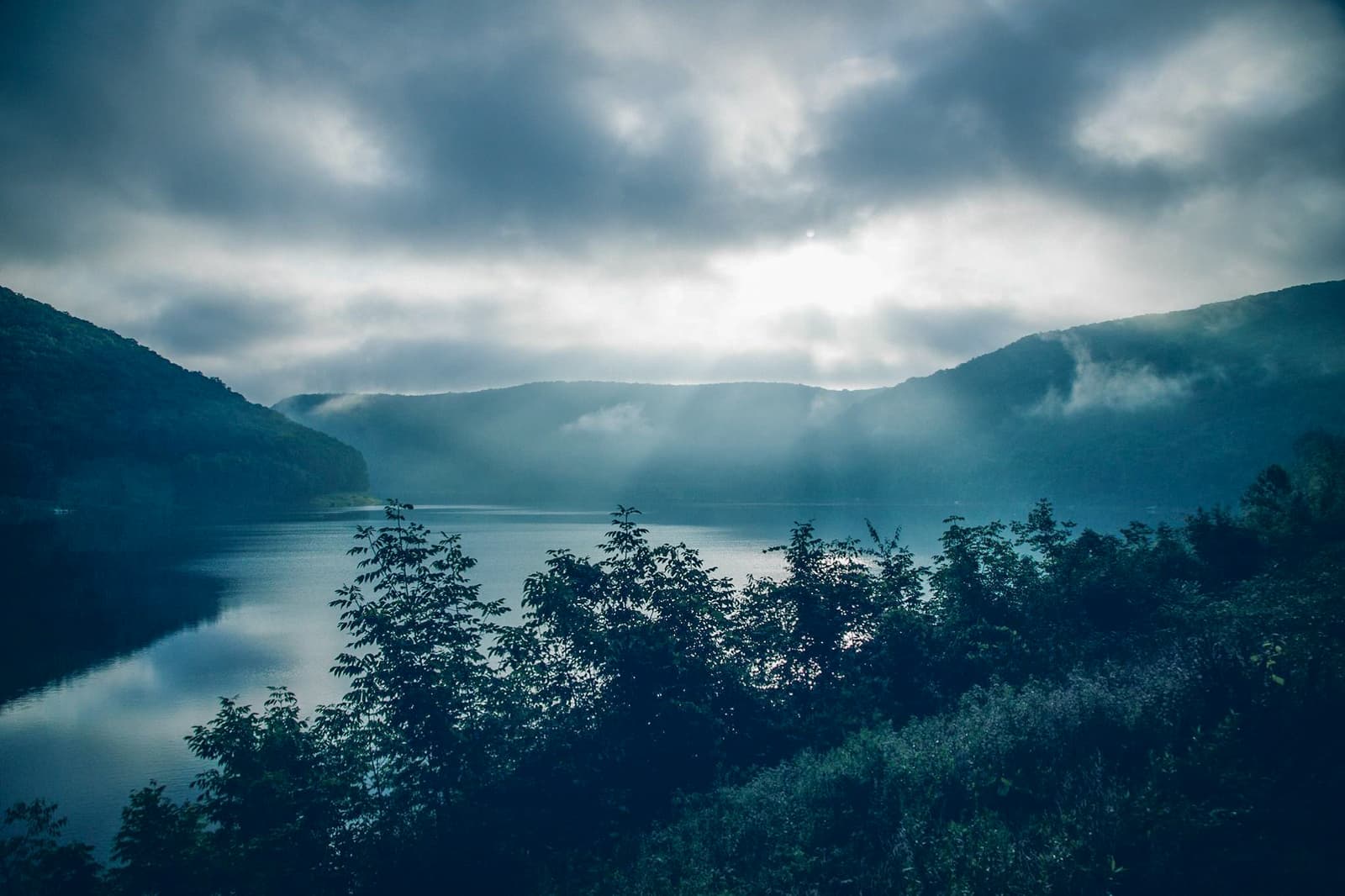 A misty lake surrounded by forested hills under a cloudy sky. Sunlight breaks through the clouds, casting soft rays onto the water and highlighting the lush greenery along the shore.