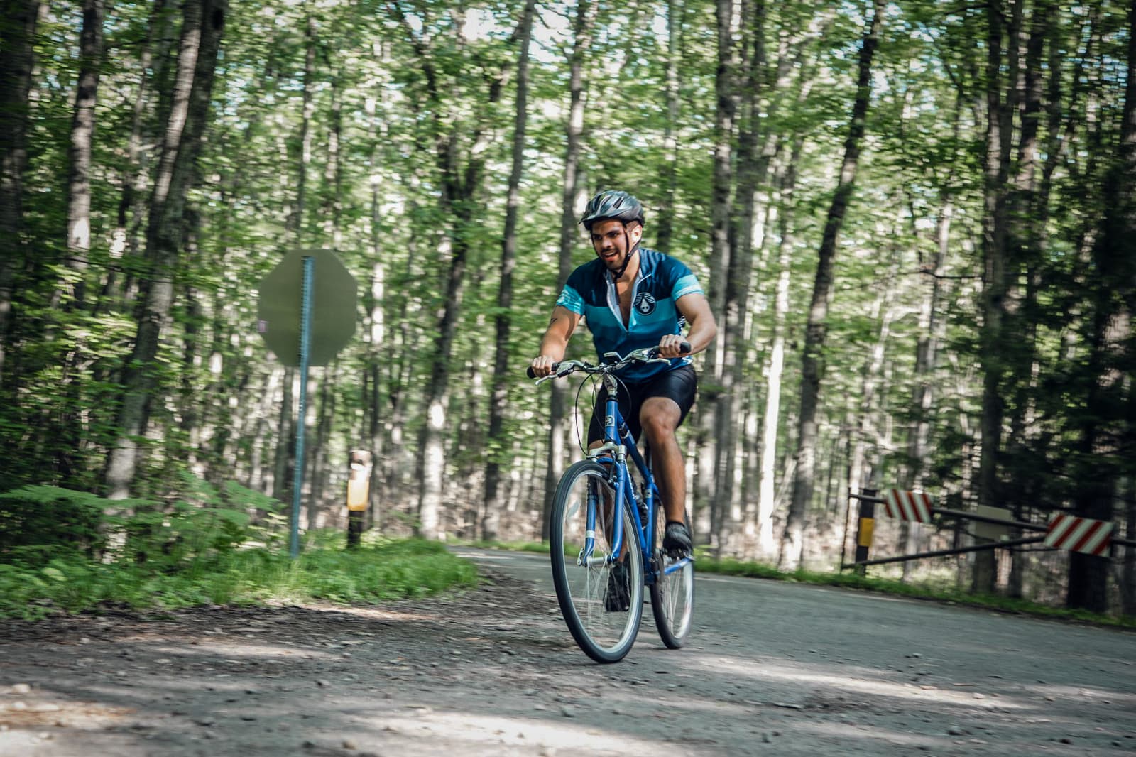 A man wearing a blue cycling jersey and helmet rides a bicycle on a forested path. Trees with green leaves surround him, and a road sign is visible in the background.