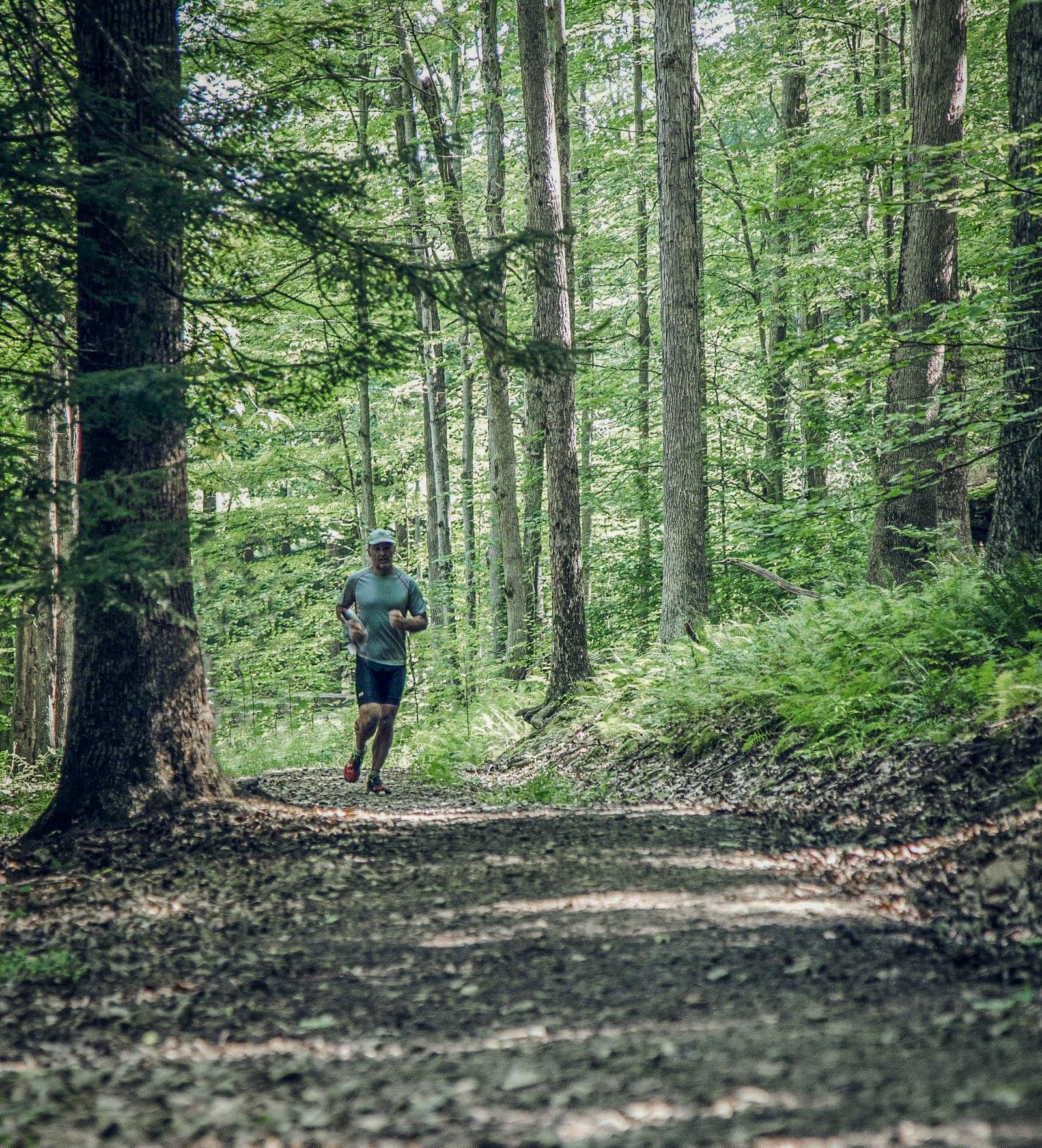 A person runs along a leaf-covered dirt trail in a lush, green forest. Sunlight filters through tall trees and dense foliage, creating a peaceful, natural atmosphere.