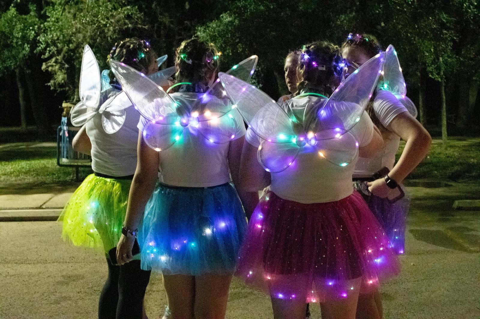 Four people wear glowing fairy wings and colorful tutus, standing together outside at night. Their outfits are lit with multicolored lights, and trees are visible in the background.