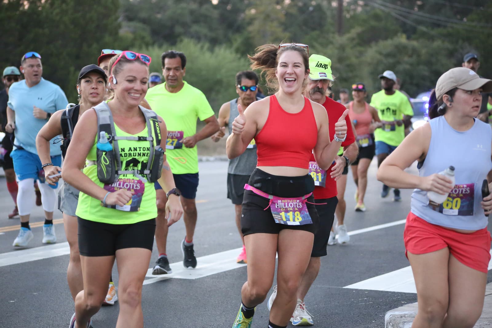 A group of runners, both men and women, participate in a race on a road. Two women in the foreground, smiling and giving thumbs up, wear running gear and race bibs. Other runners are seen behind them.