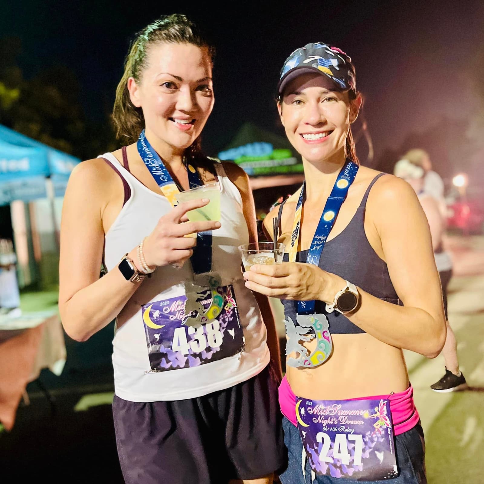 Two women wearing race bibs and medals smile while holding drinks at an outdoor event. Both wear athletic outfits and appear happy, suggesting they just finished a race. Tents and lights are visible in the background.