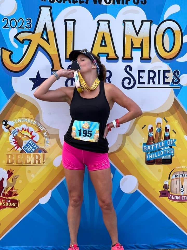 A woman wearing a black tank top, pink shorts, and a race bib numbered 195 kisses a medal in front of a colorful "2023 Alamo Beer Series" backdrop, celebrating her achievement.