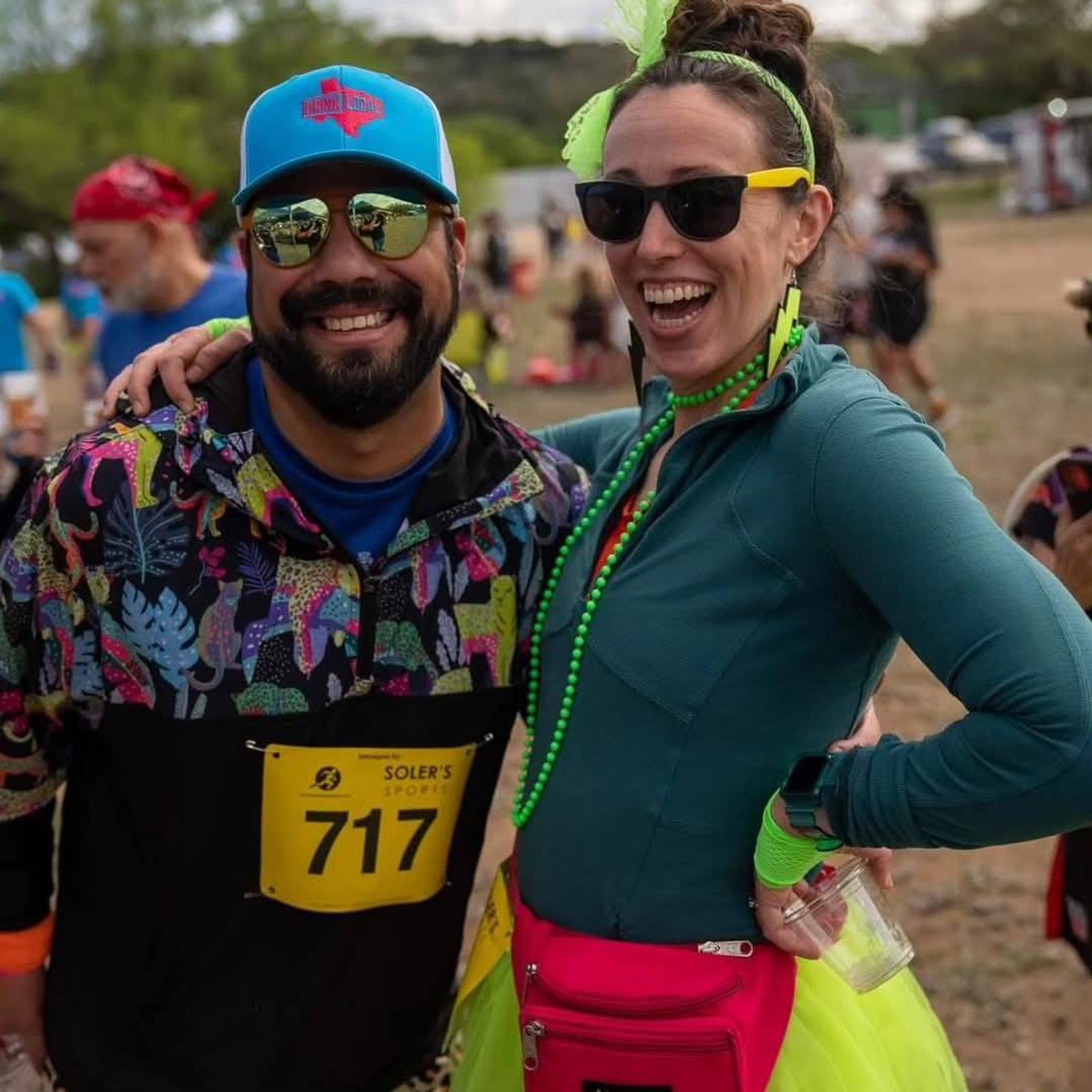 Two people at an outdoor event, smiling at the camera. One wears a blue hat, sunglasses, and a colorful jacket with a race bib. The other wears neon accessories, a tutu, sunglasses, and a bright pink fanny pack.