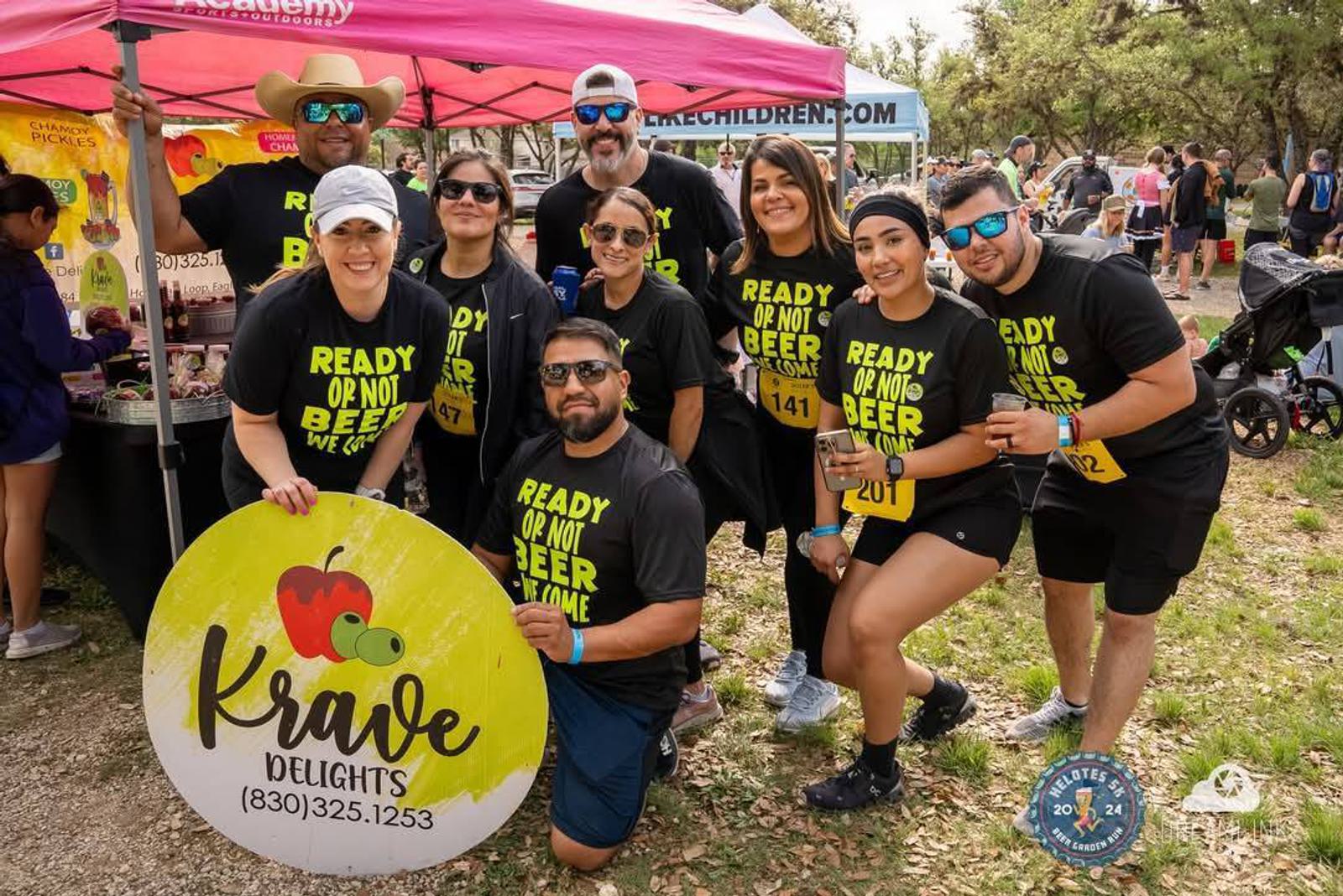 A group of nine smiling people pose outdoors at an event, wearing matching black "Ready or not, beer I come" shirts and race bibs, holding a “Krave Delights” sign in front of a pink tent. Trees and other participants are in the background.