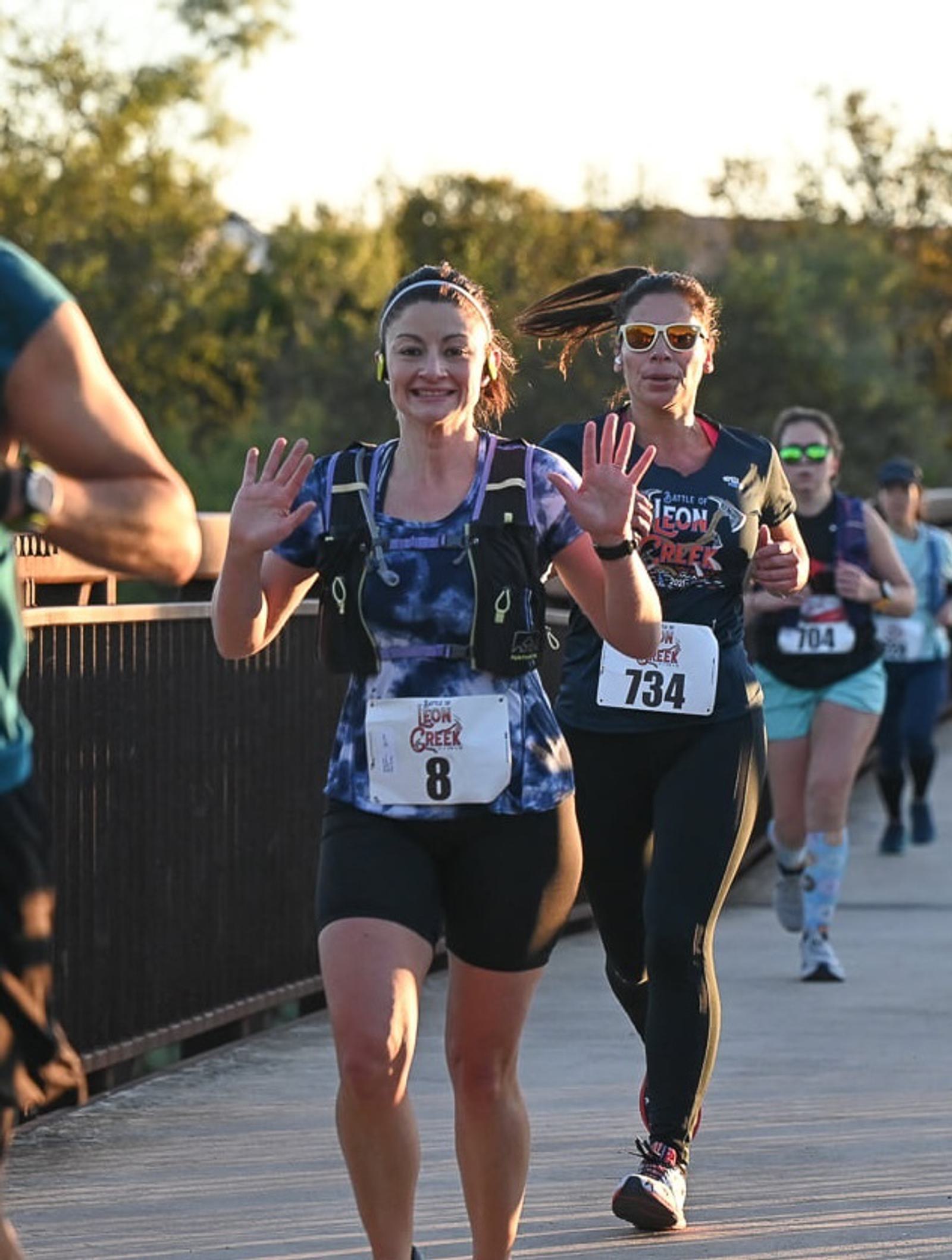 A woman running a race on a bridge smiles and waves at the camera, with other runners behind her. She wears a numbered race bib and athletic gear. The sun is shining and trees are in the background.