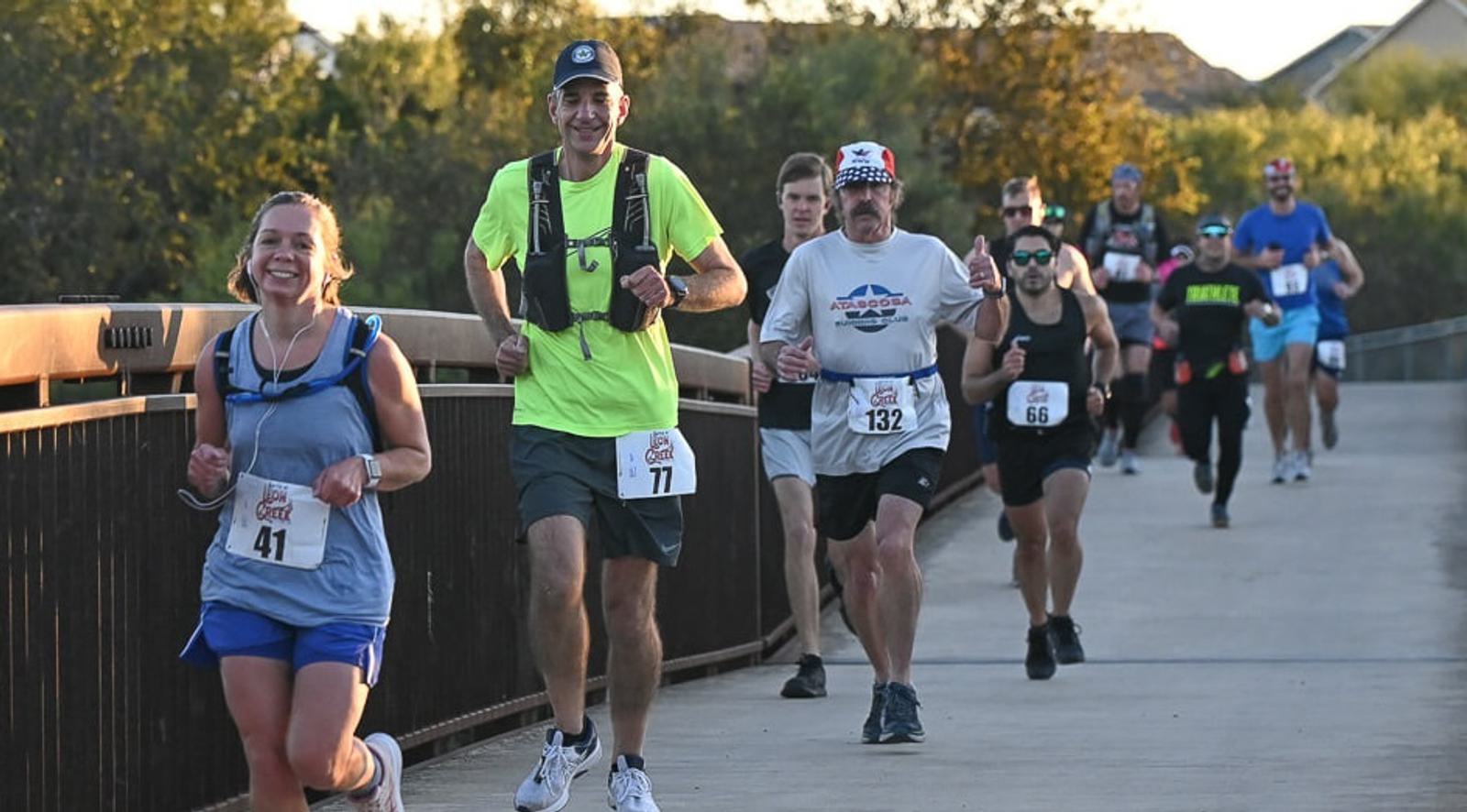 A group of runners with race bibs jogs across a bridge during a race on a sunny day. Some are smiling, and others look focused. Trees and houses are visible in the background.