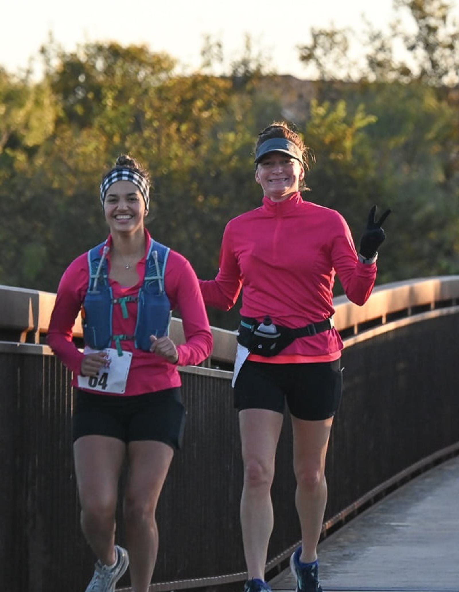 Two women running outdoors on a bridge, both wearing pink tops and black shorts. One woman is smiling at the camera, while the other flashes a peace sign. Trees are visible in the background.