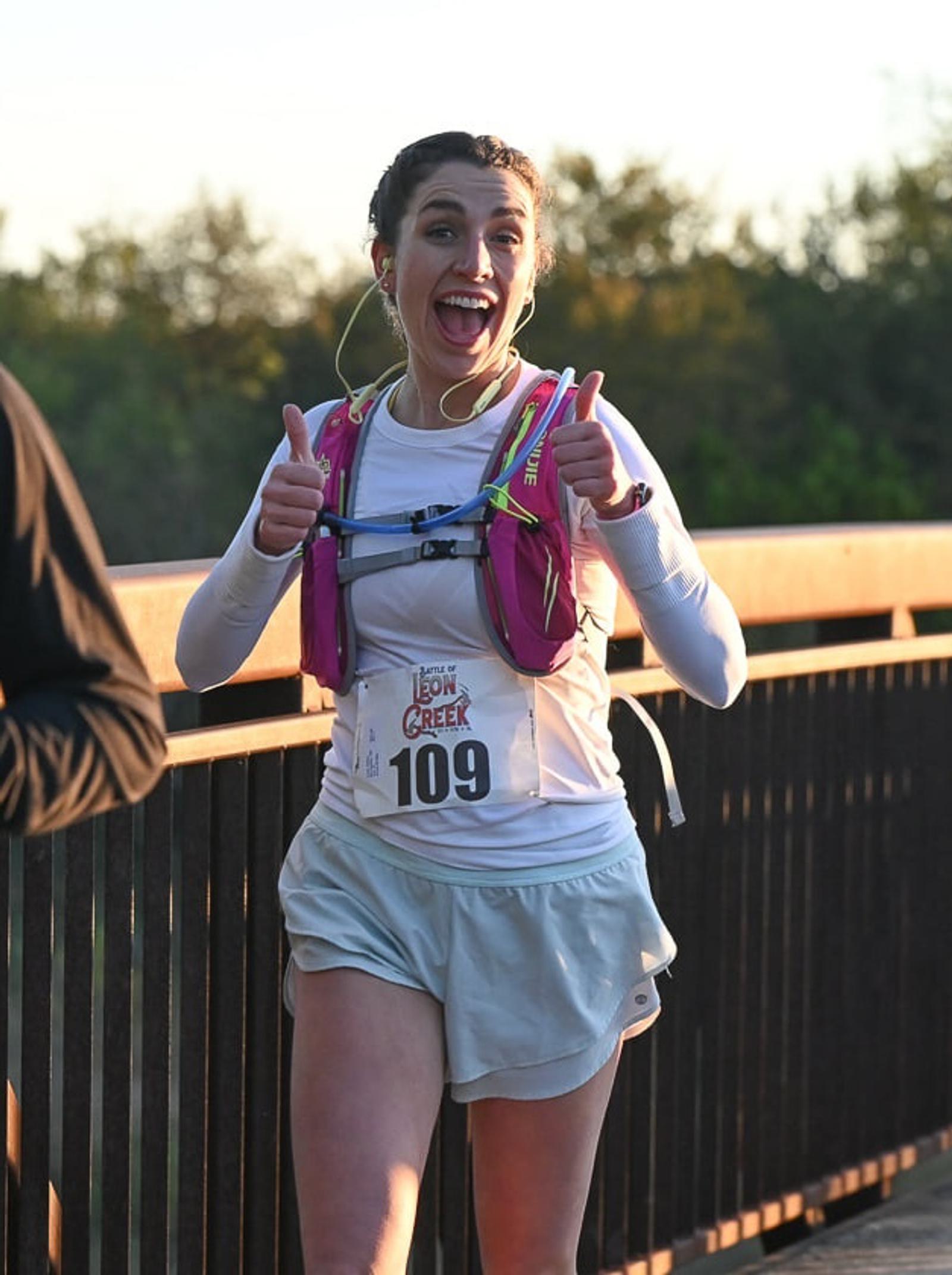 A smiling woman wearing a race bib numbered 109 gives two thumbs up while running outdoors on a bridge, with trees in the background. She wears headphones, a white shirt, and a pink hydration pack.