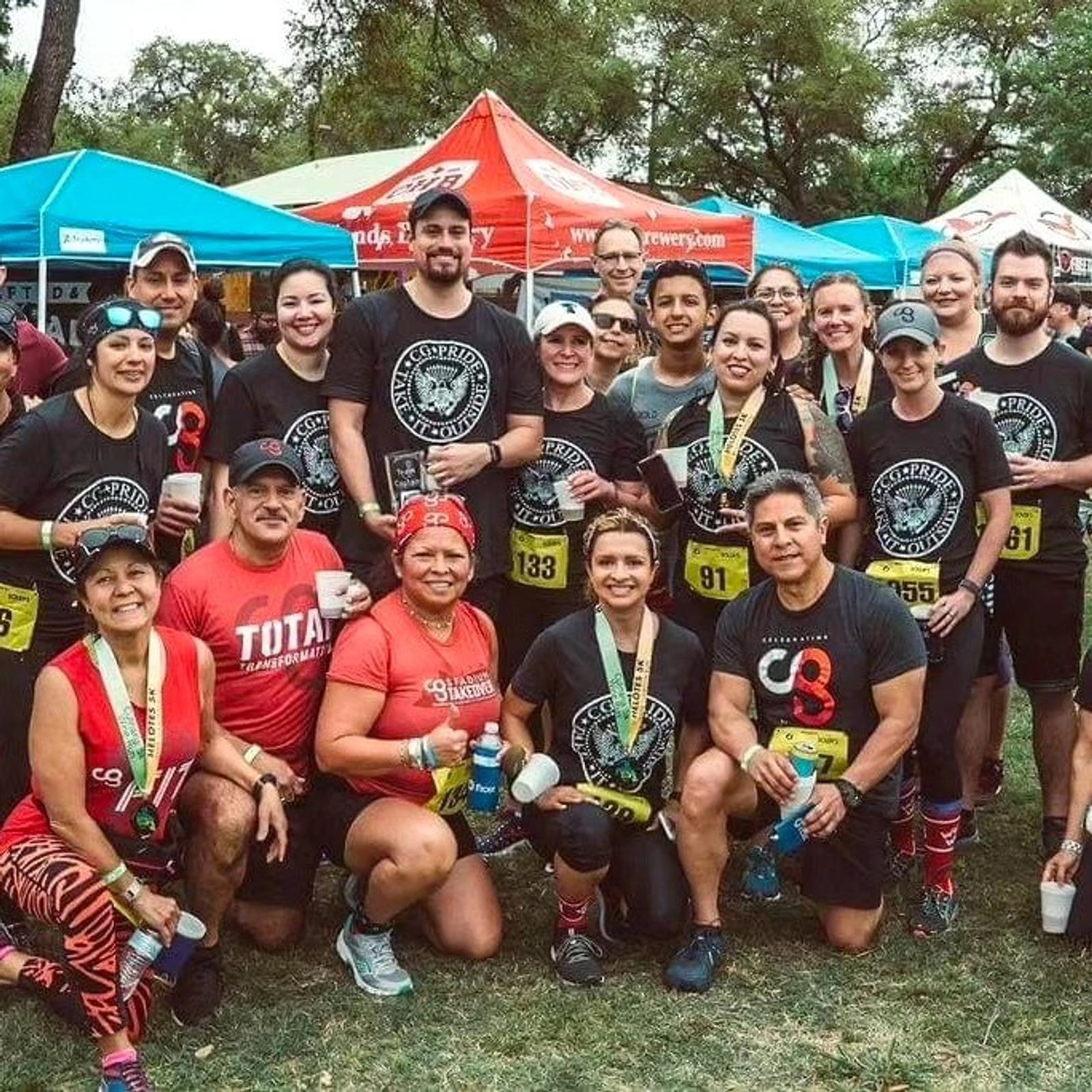 A group of runners wearing race bibs and matching shirts pose and smile together outdoors at a race event, with colorful tents and trees in the background.