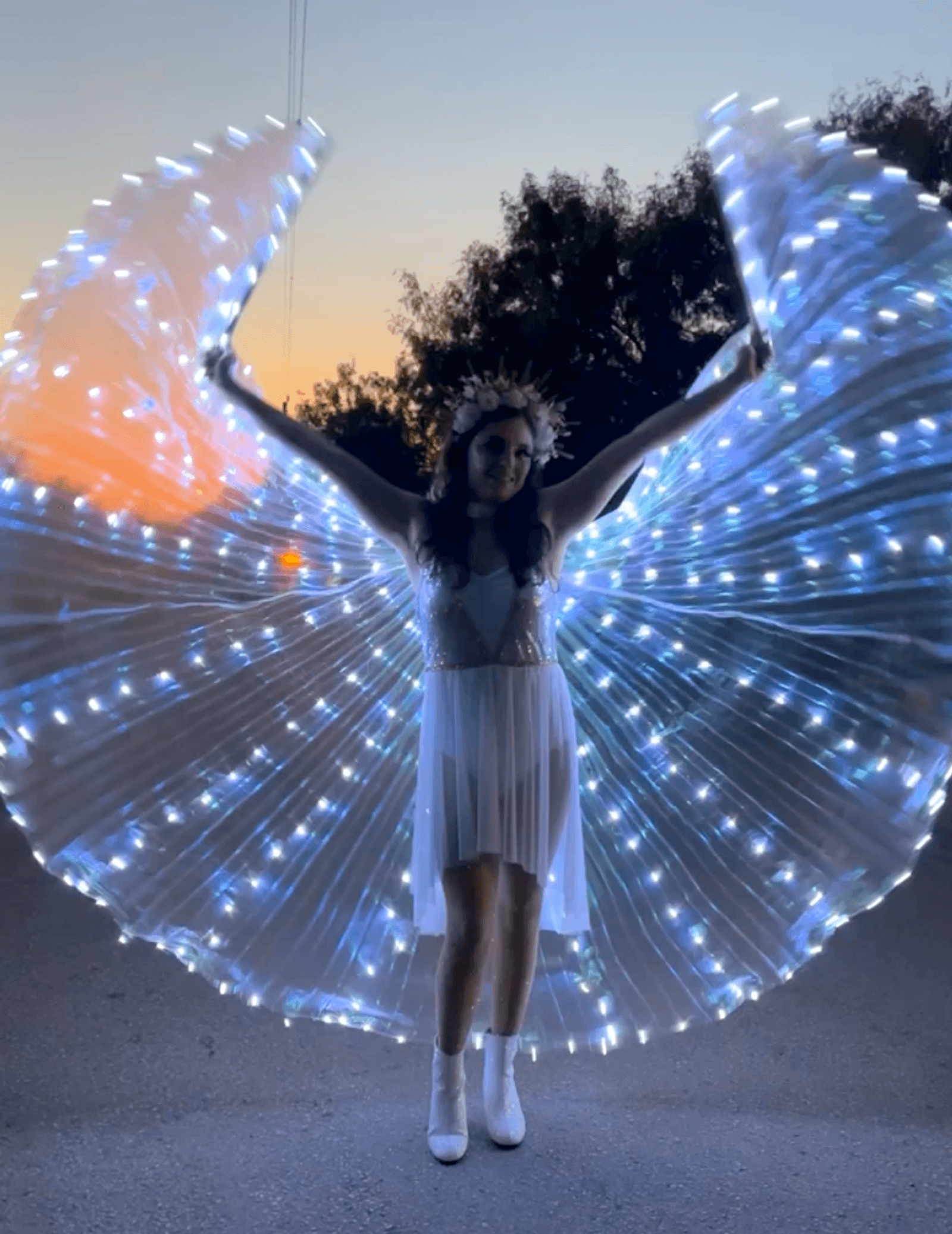 A person in a white dress and boots stands outdoors at dusk, wearing illuminated wings made of glowing white lights spread wide. Trees and the fading sunset sky are visible in the background.