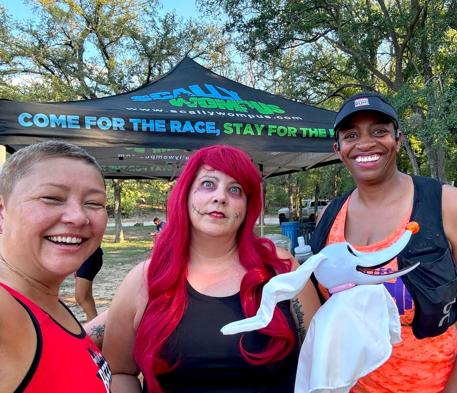 Three people pose and smile outdoors in front of a tent that says, "Come for the race, stay for the party." One person wears a red wig and face paint, another holds a white costume prop, and trees are visible in the background.