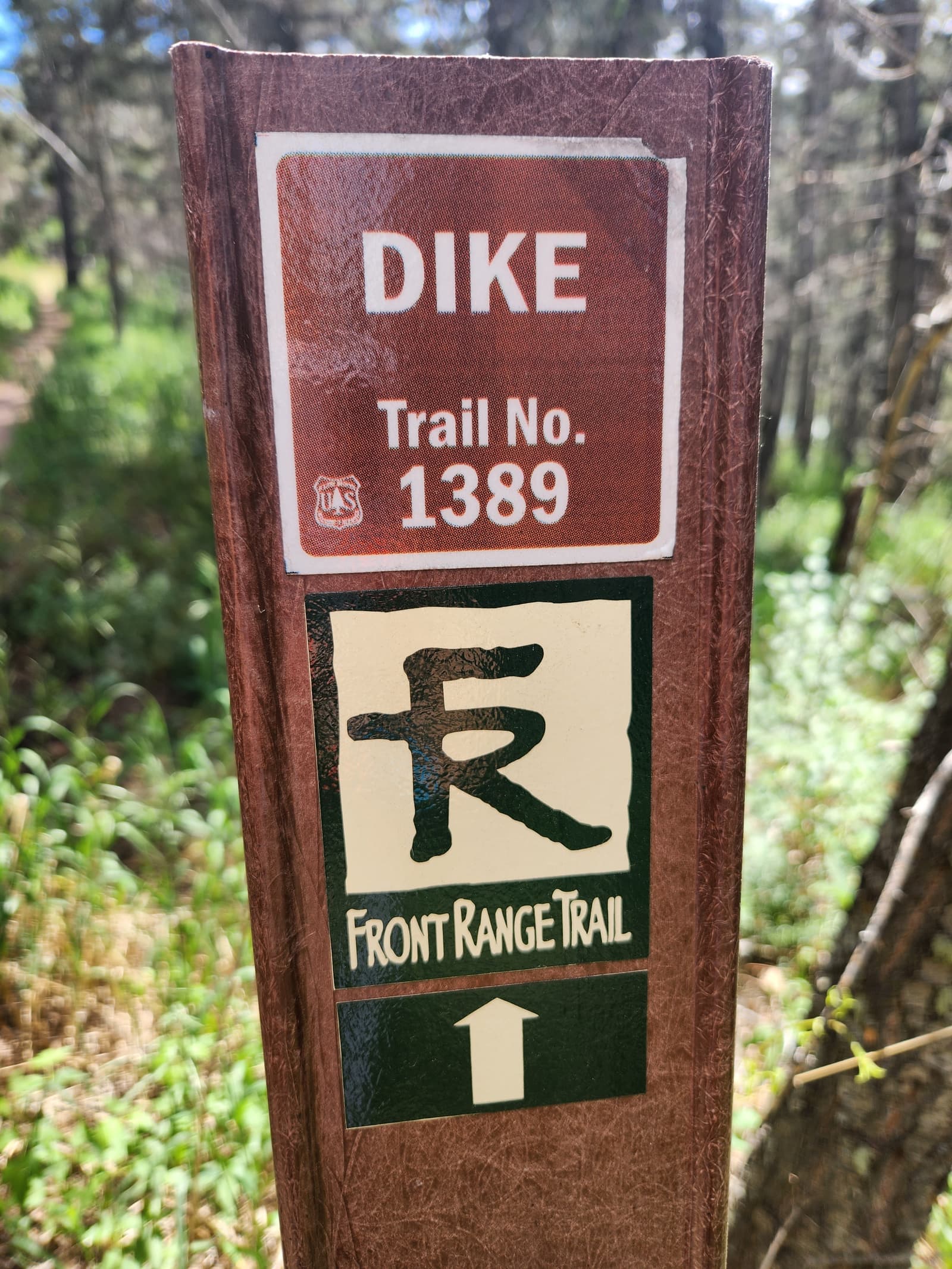 A wooden trail marker with signs reading "DIKE Trail No. 1389" and "Front Range Trail" stands beside a forested path, with sunlight filtering through the trees.