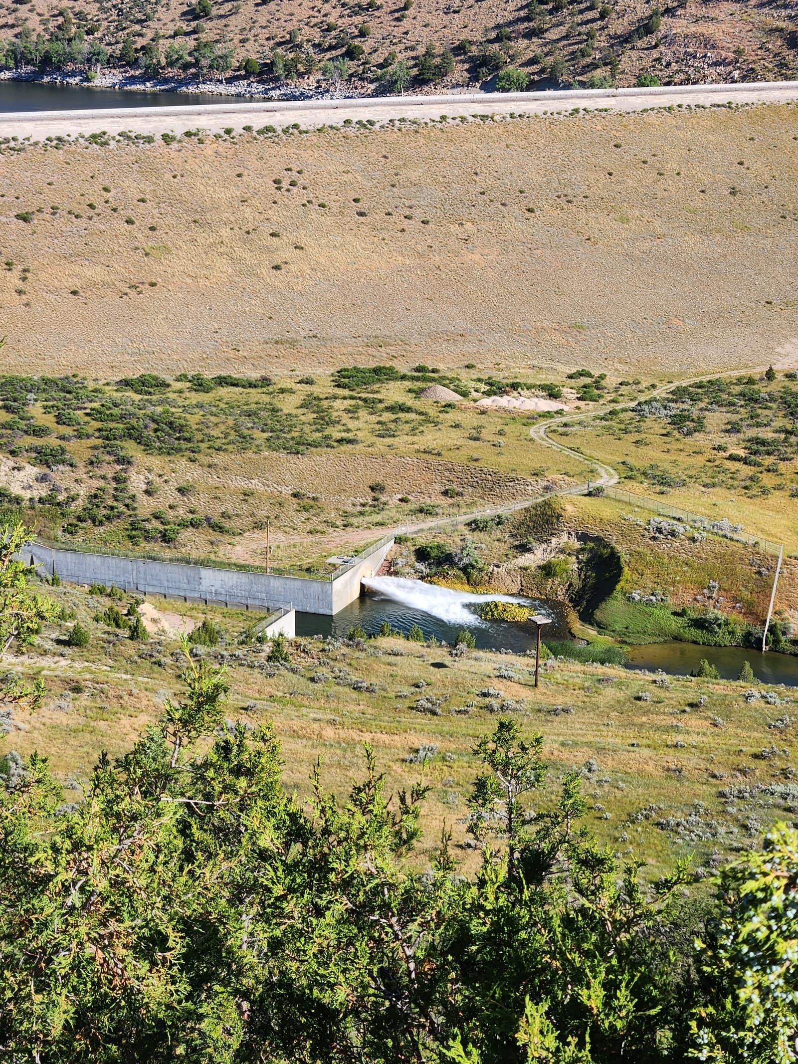 A small concrete dam structure stands in a grassy, hilly landscape with bushes and a winding path nearby. A large reservoir and gravel road stretch across the top of the image.
