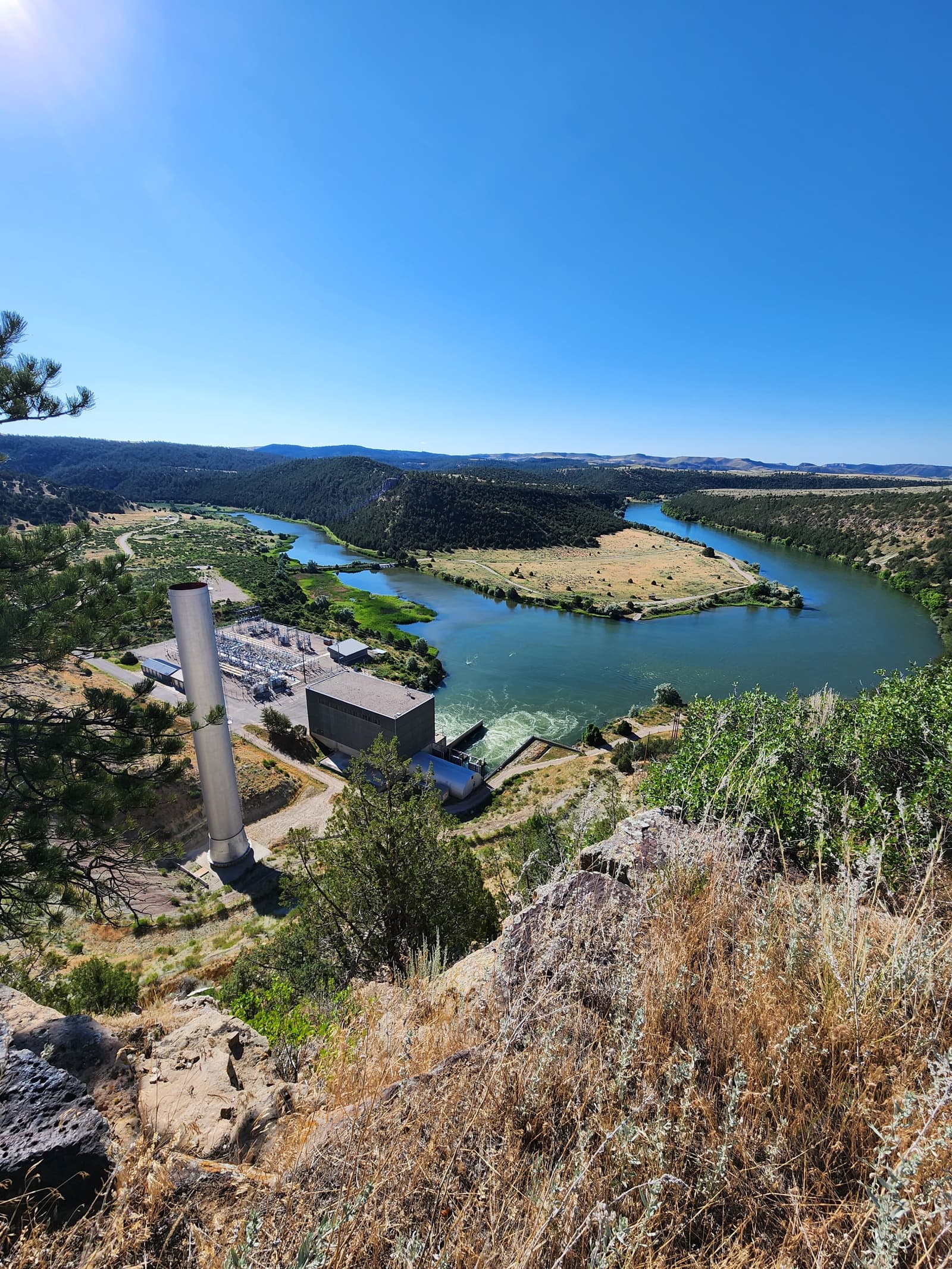 View from a rocky hill overlooking a winding river, green fields, and a small industrial facility with two tall chimneys; forests and distant hills are visible under a clear blue sky.