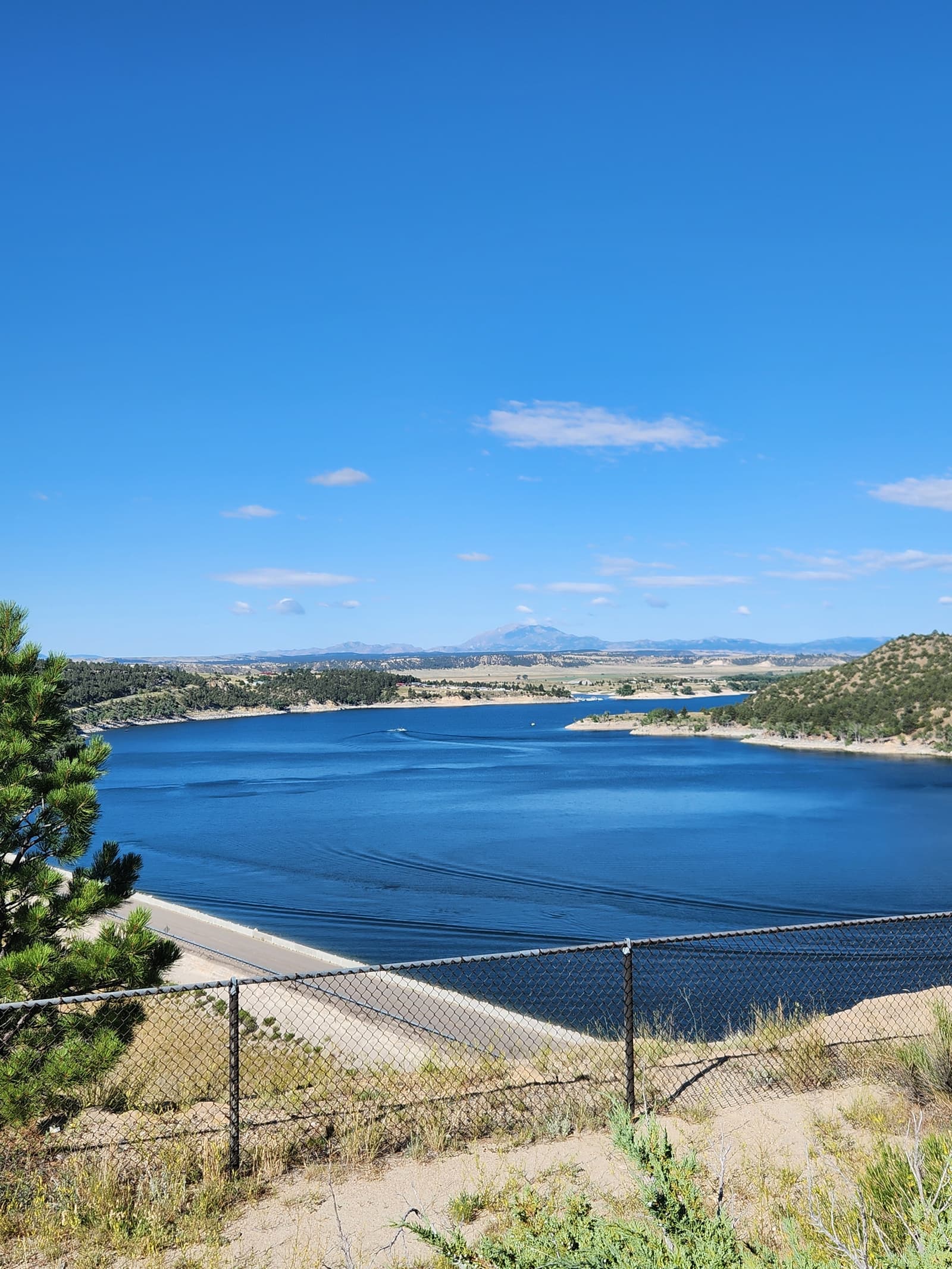 A large blue reservoir surrounded by rolling hills and trees under a clear blue sky, viewed from behind a chain-link fence with some greenery in the foreground.